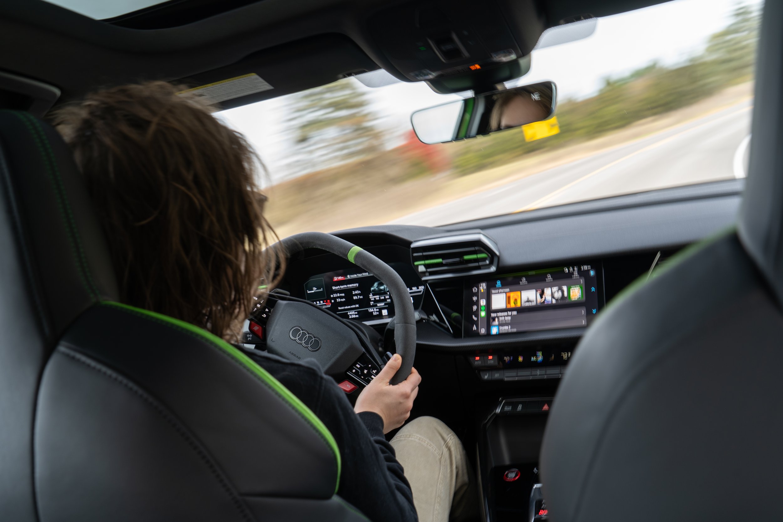 Person with curly hair driving an Audi vehicle with a touchscreen dashboard on a road with trees in the background.