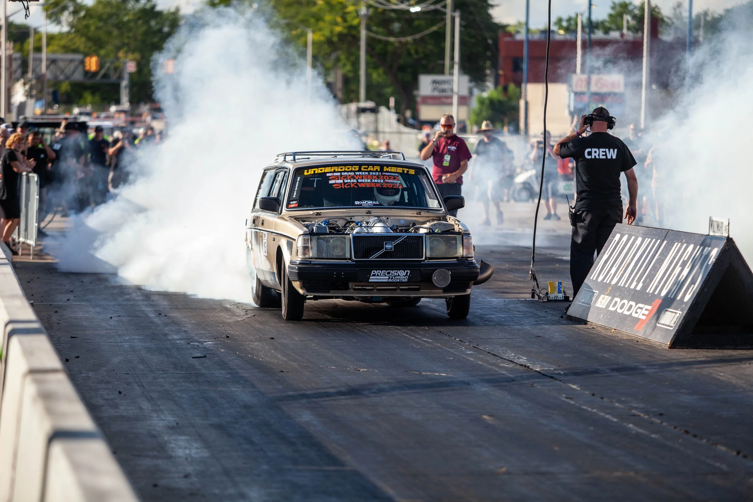 A drag racing car on the starting line with smoke behind it, crew member wearing a black shirt with 'CREW' on the back, and spectators observing.