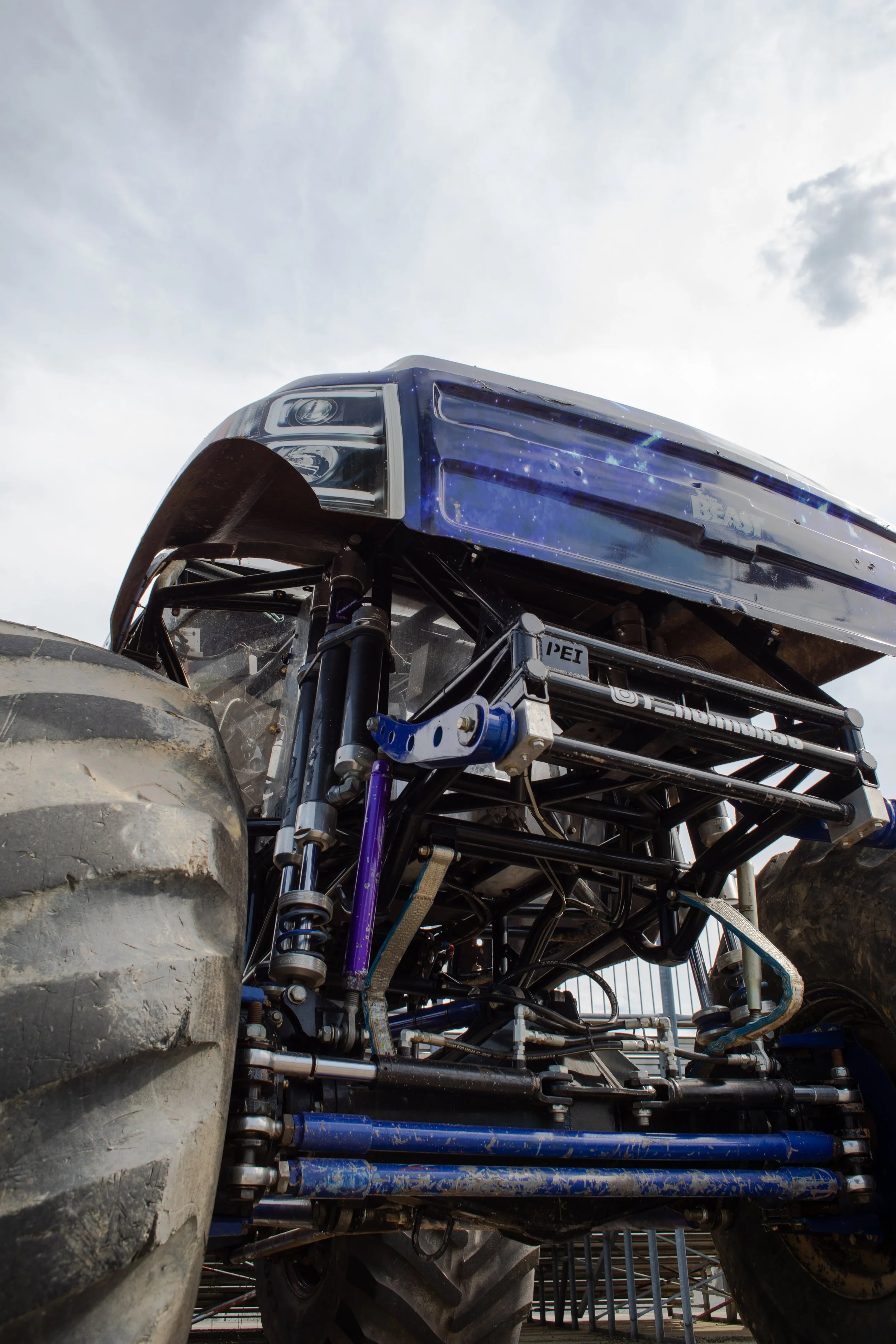 Close-up view of a large off-road vehicle with exposed suspension, tires, and a blue body with galaxy-like artwork, under a cloudy sky.