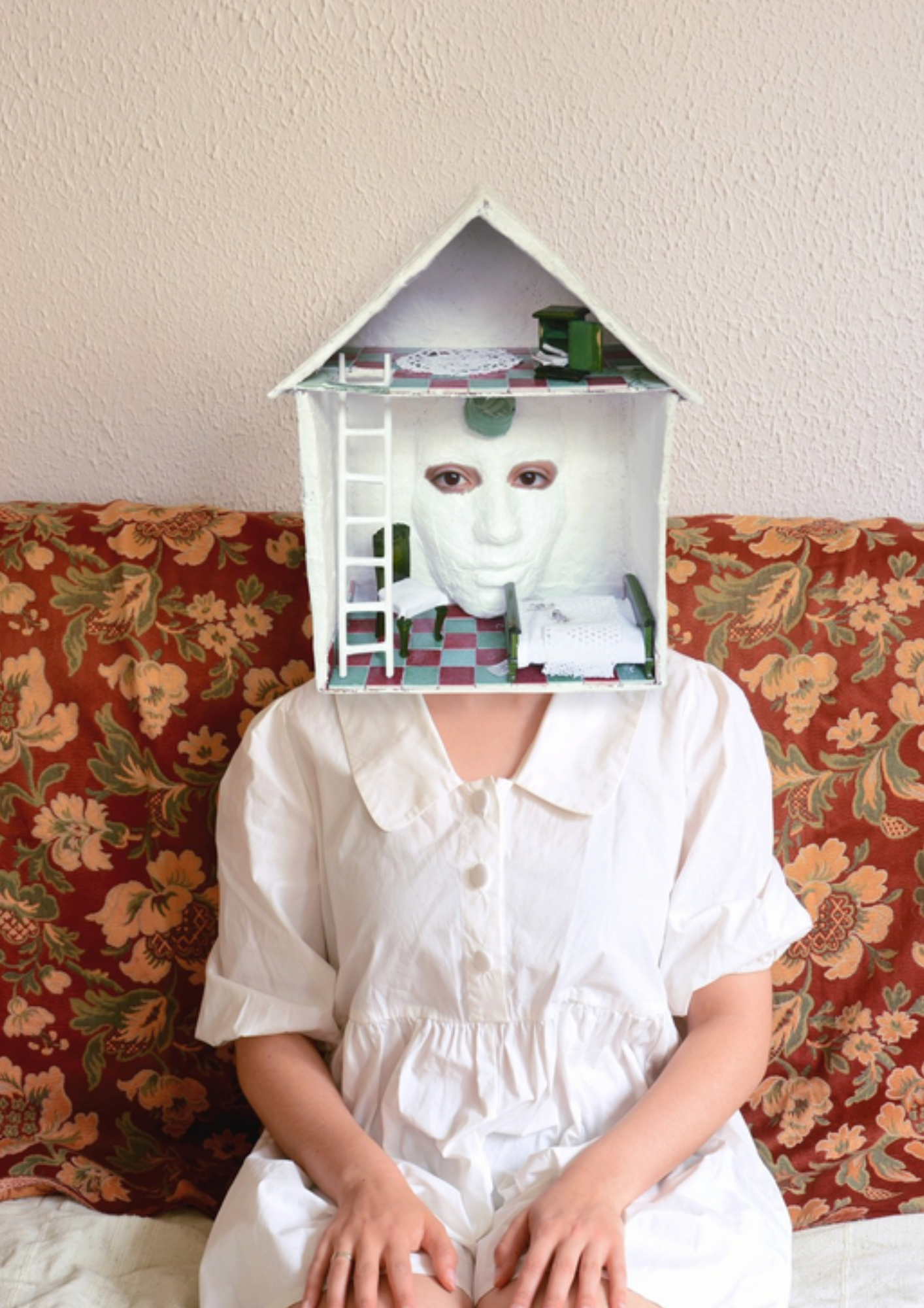 Une personne assise sur un canapé à motifs floraux, portant une chemise blanche et un masque en forme de visage humain, avec une maison miniature sur sa tête.