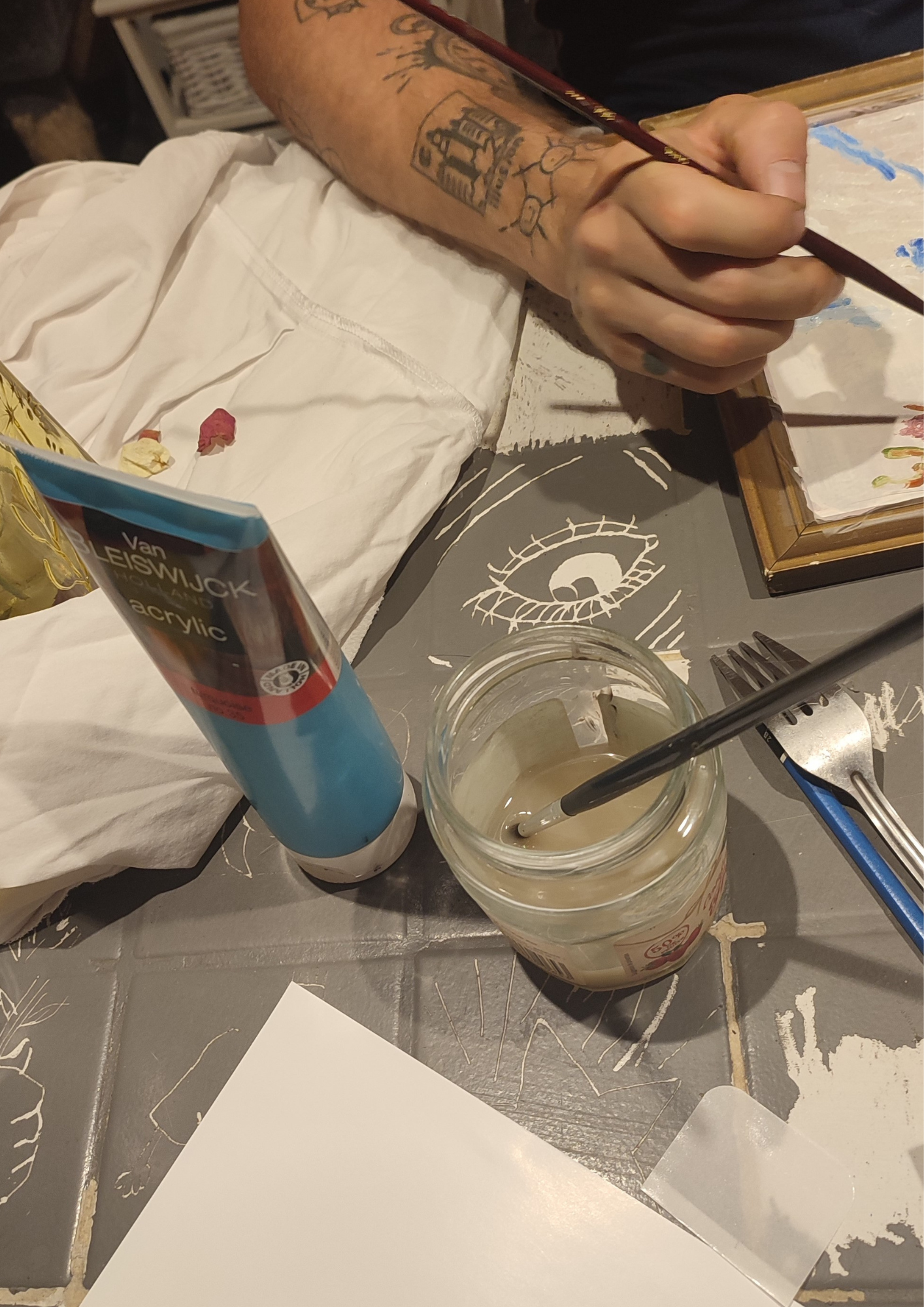 Une main tenant un pinceau, en train de peindre ou de créer de l'art, avec des tubes de peinture acrylique, un pot de pinceaux dans un cerveau dessinée avec de la peinture blanche, et des éléments artistiques autour.