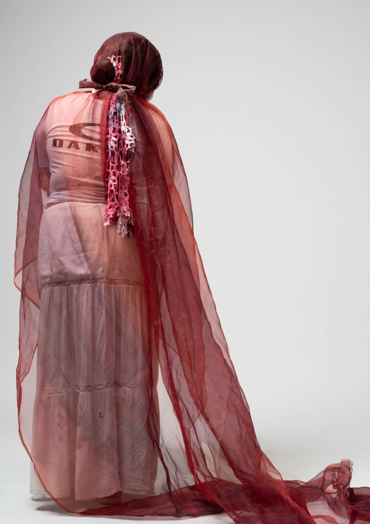 Une personne vue de dos, portant une robe longue et transparente, avec un châle rouge, sur un fond blanc.