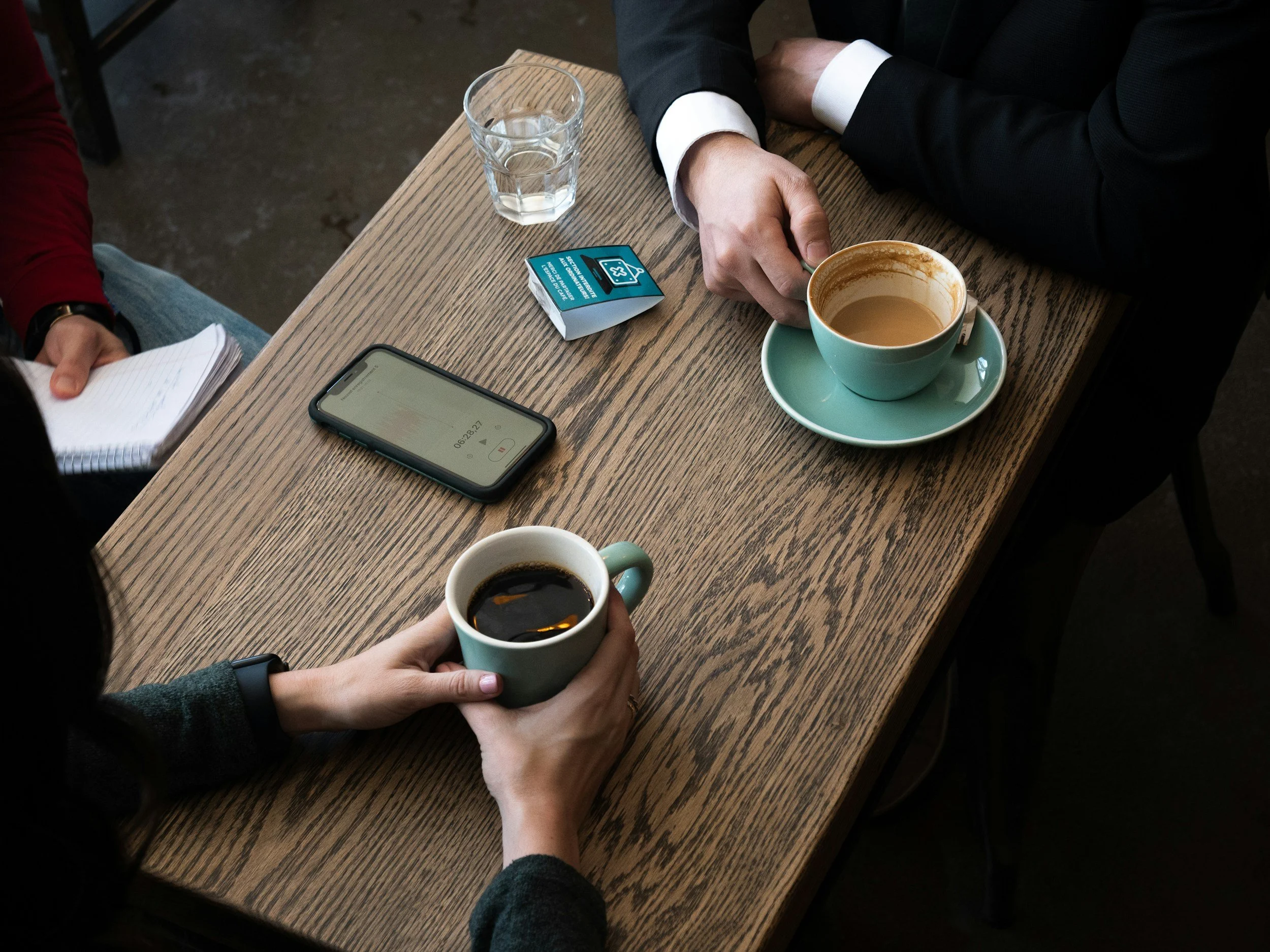 People sitting at a wooden table in a café, one person holding a cup of black coffee, another with a cup of coffee with cream or milk, with a glass of water, a smartphone, a notepad, a tampon box, and a cup of coffee on the table.