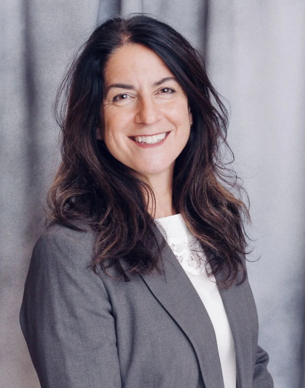 A woman with long dark hair, wearing a gray blazer and white top, smiling at the camera against a plain gray background.