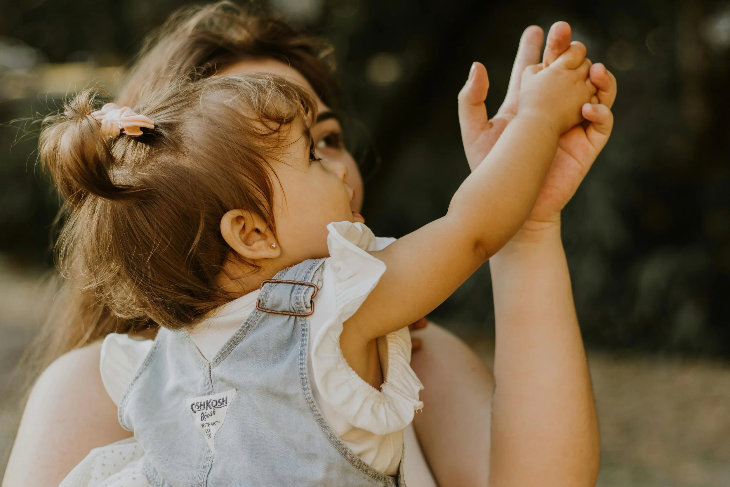A woman holding a young girl in an outdoor setting. The girl has her hands clasped with the woman's hands, with her hair in a small ponytail secured with a pink hairclip. The girl appears to be looking up and reaching out with her hands.
