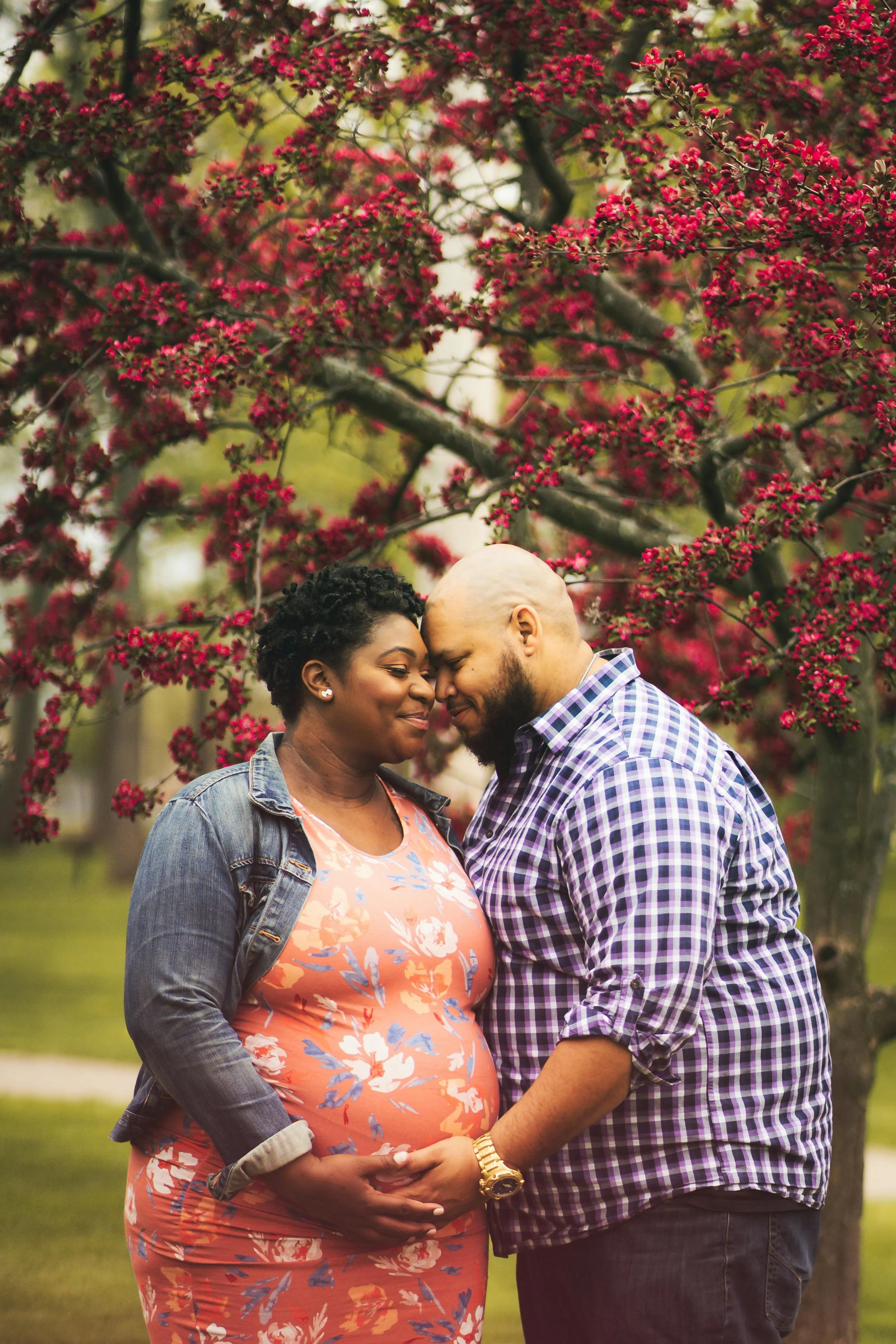 Two expecting parents under a flowering tree, both with their hands on the gestational parent's belly.