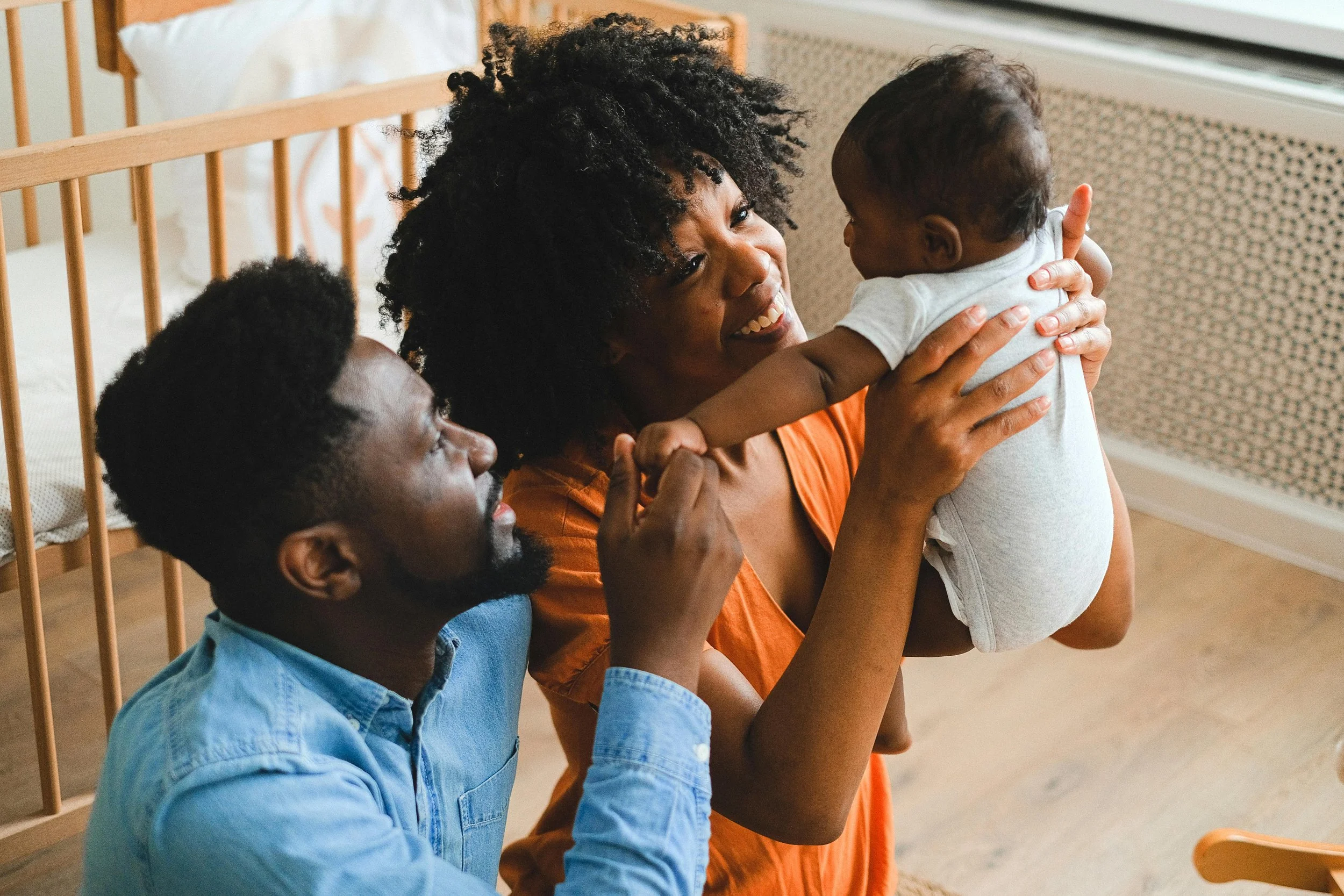 A woman holding a baby up, with a man close by, smiling and engaging with the baby in a bright room.