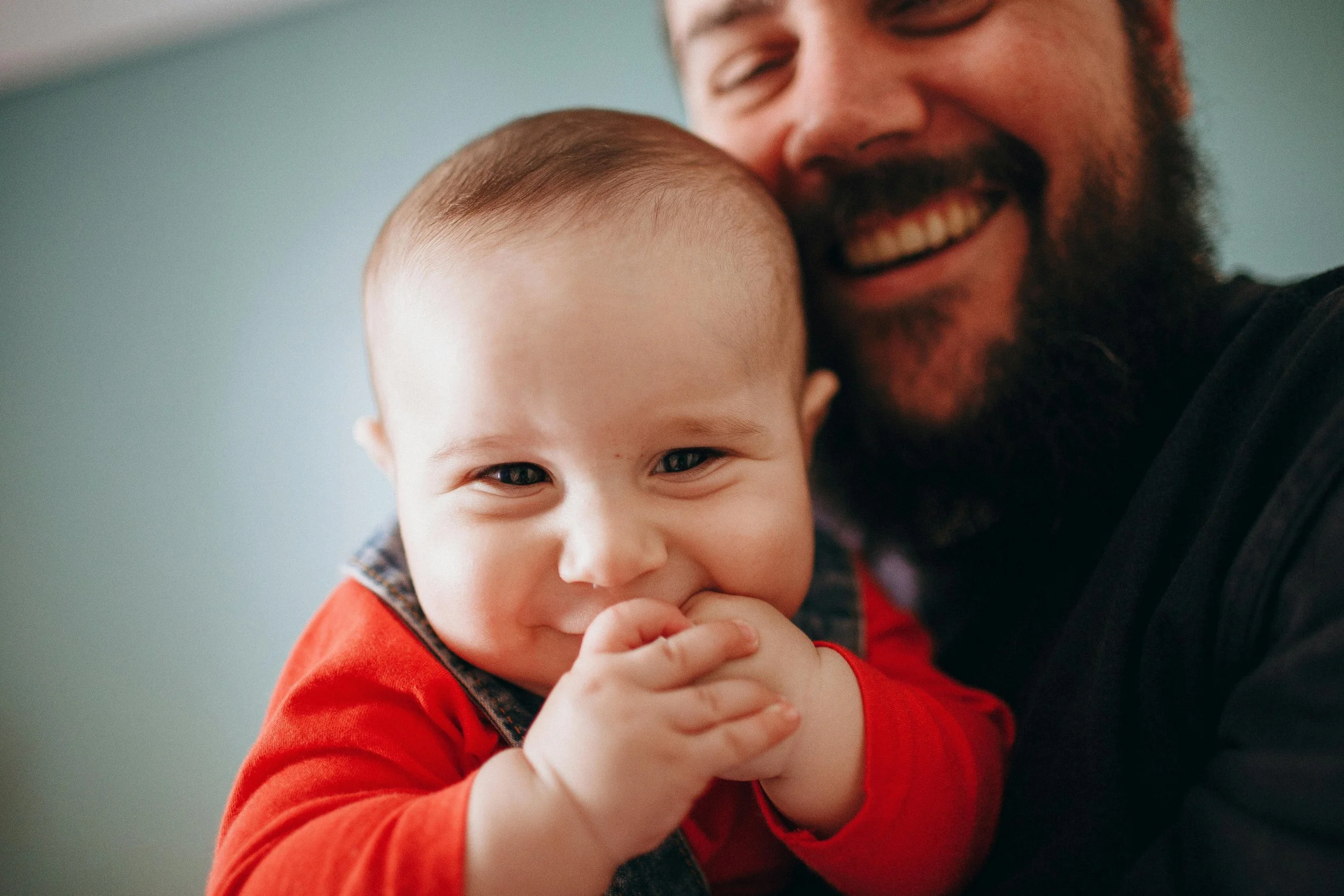 A smiling baby with hands near mouth, being held closely by a man with a beard and a big smile, both enjoying a happy moment together.