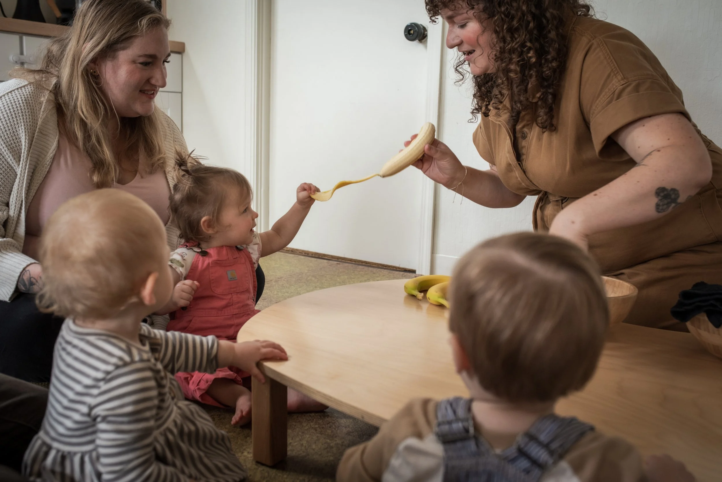 A toddler in pink overalls helps to peel a ripe banana held by a curly haired educator in a brown jumpsuit.