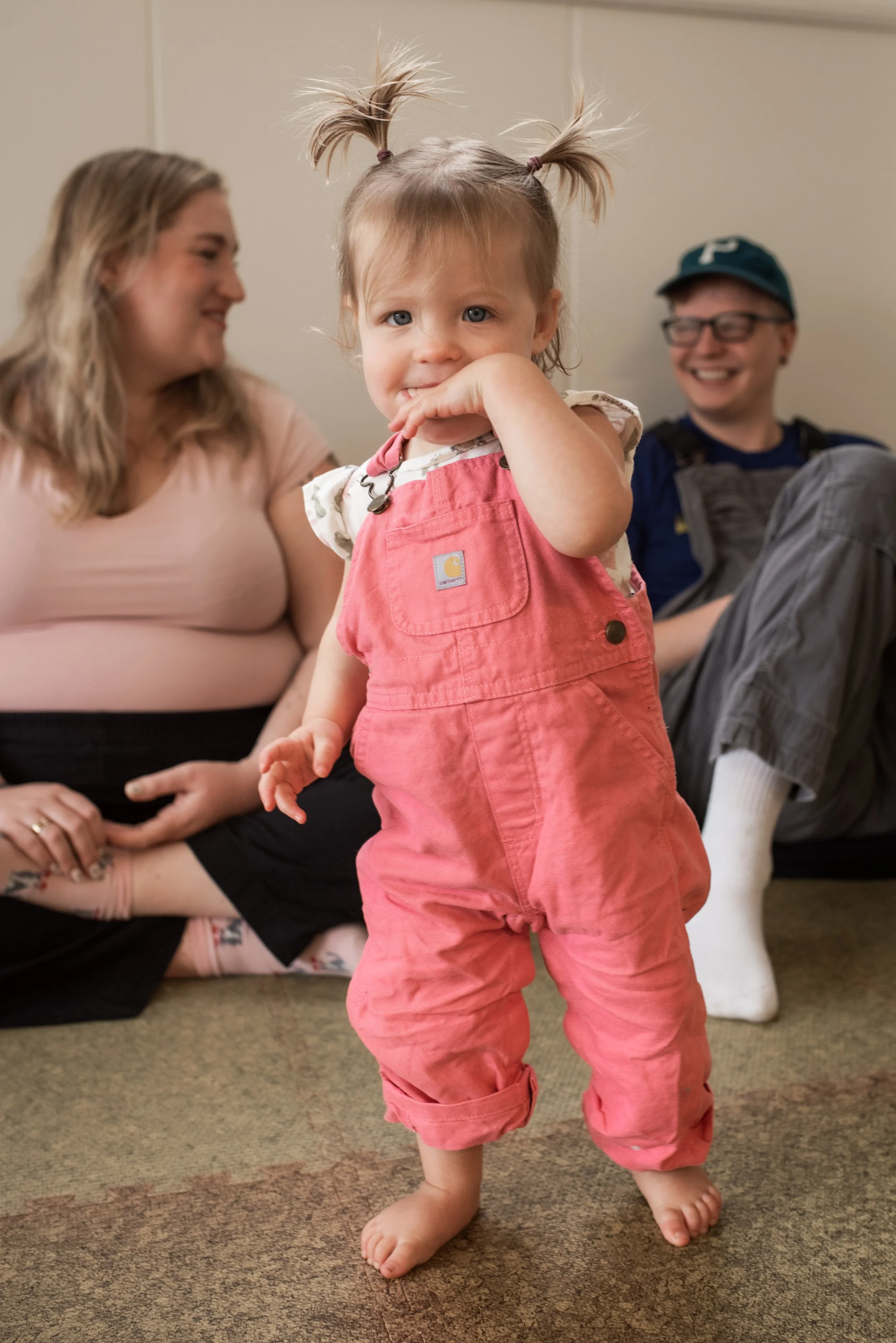 A toddler with pigtails and pink overalls is in the foreground. Behind her are one parent with blond long hair and one with glasses and a green ballcap smiling at each other.