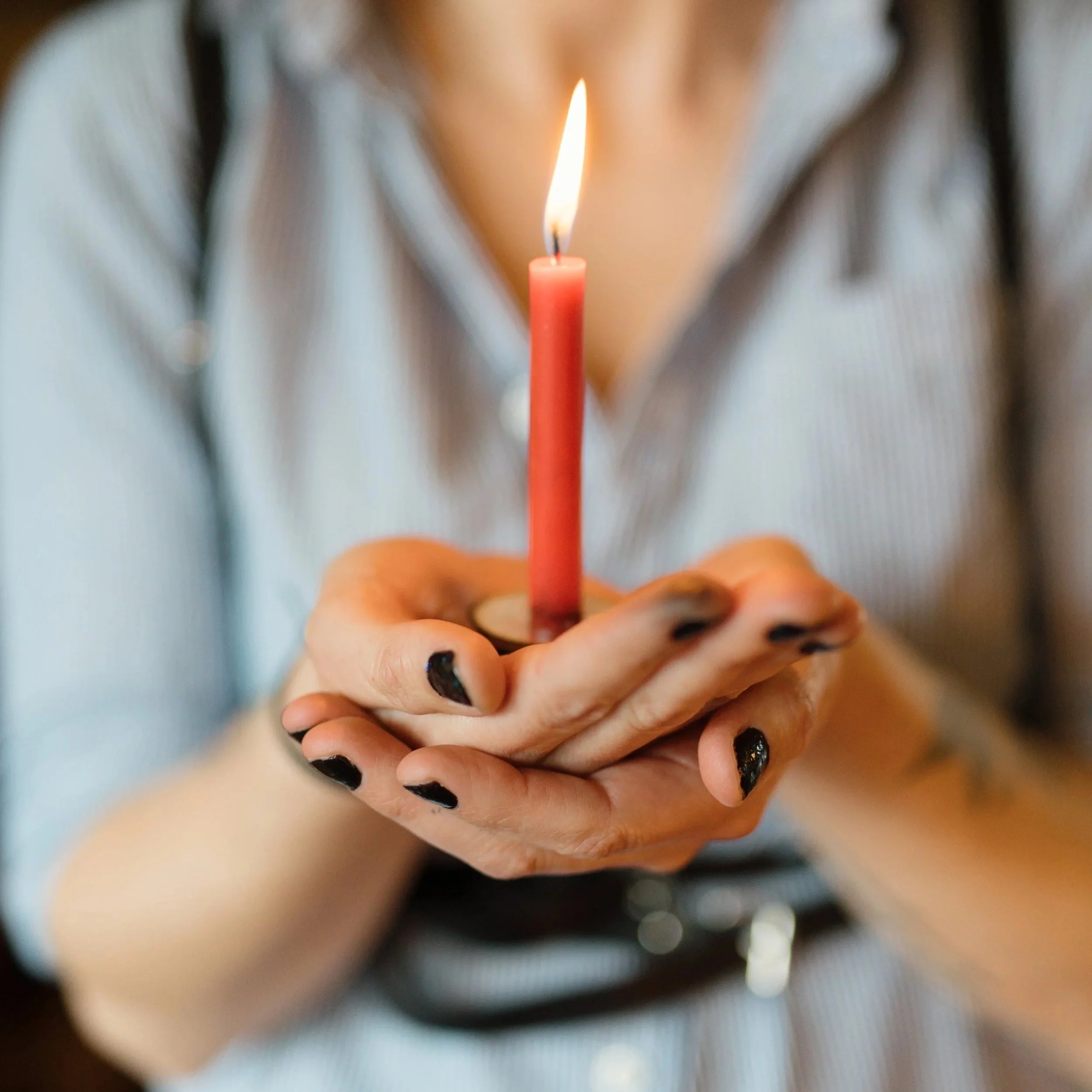 A person holding a lit birthday candle with black nail polish on their fingernails.