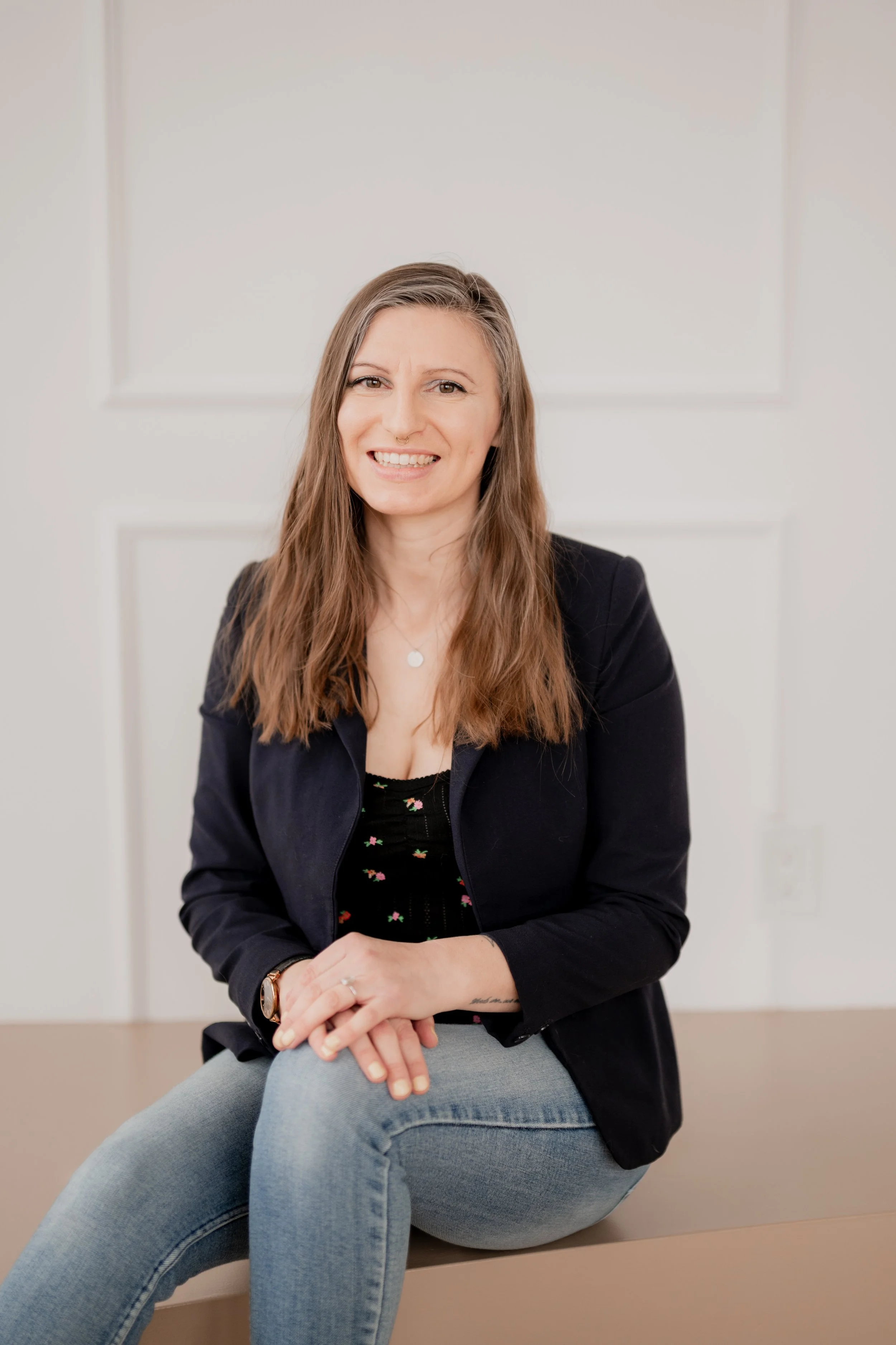 A woman with long light brown hair, smiling, sitting on a beige surface, wearing a black blazer, a floral top, and light blue jeans, in a bright room with white walls.