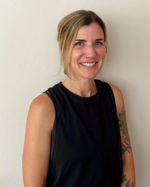 A woman with blonde hair in a ponytail, wearing a black sleeveless top, smiling, standing against a white wall, revealing tattoos on her left arm.
