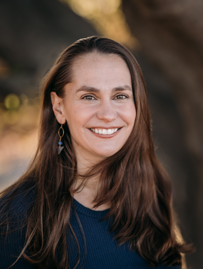 Portrait of a woman with long brown hair, smiling, wearing a navy top and earrings, outdoors with a blurred rocky background.