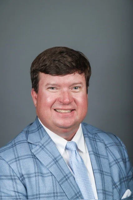 Man in a blue plaid suit jacket and white shirt with a light blue tie, smiling at the camera against a plain gray background.