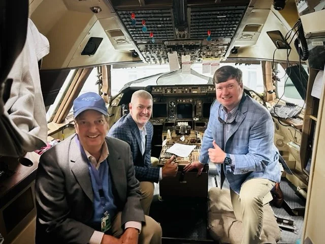 Three men sitting inside an airplane cockpit, smiling and giving thumbs up.