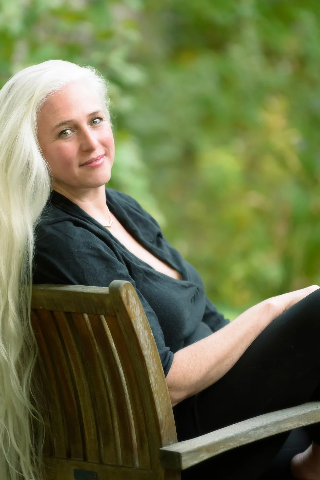 older woman with long white hair smiling gently while wearing black and seated outdoors in a teak chair