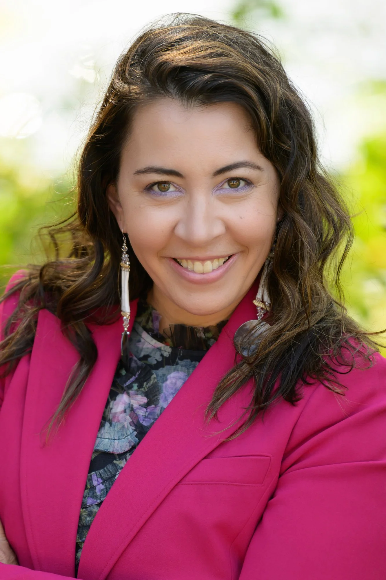 brunette woman wearing a colorful blouse and pink blazer and dangle earrings smiles brightly