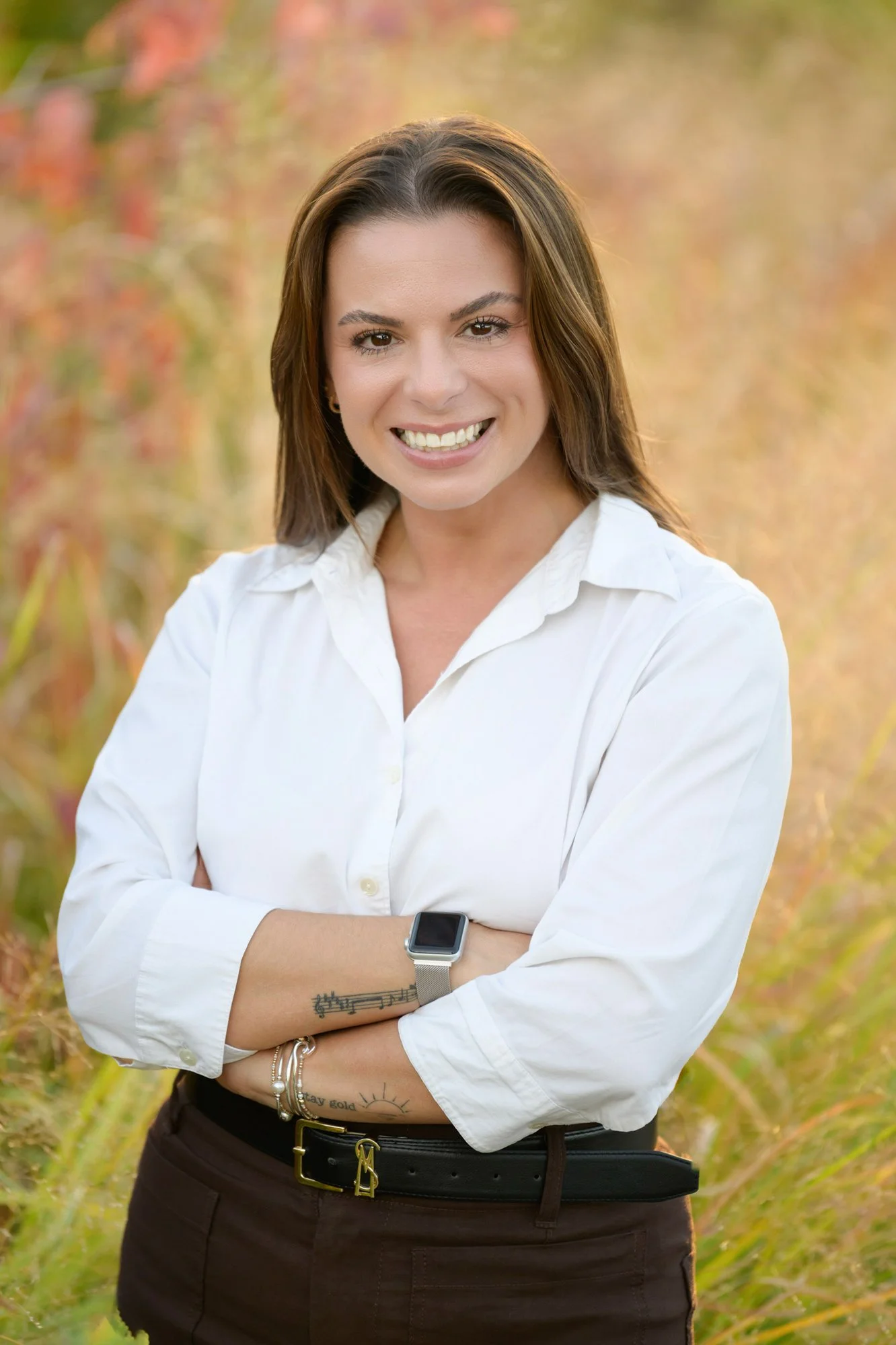 smiling brunette woman in white shirt wearing an apple watch, arms crossed with a meadow behind her