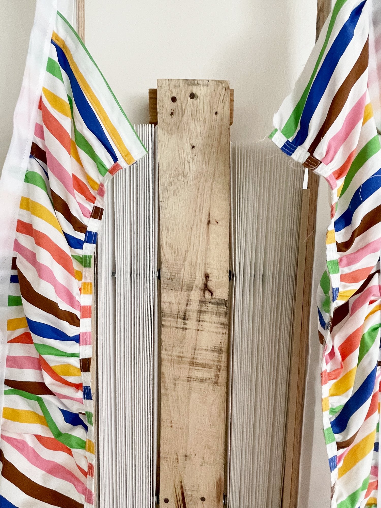 Untitled (Pride Face-hugger), 2025, 78h x 63w x 9d - salvaged wood pallets, poplar wood trim, recycled polyester white backdrop, Target Pride collection 2024 sun dress, Ikea metal white wall shelves and white metal mesh, string, threaded rod, wood gl