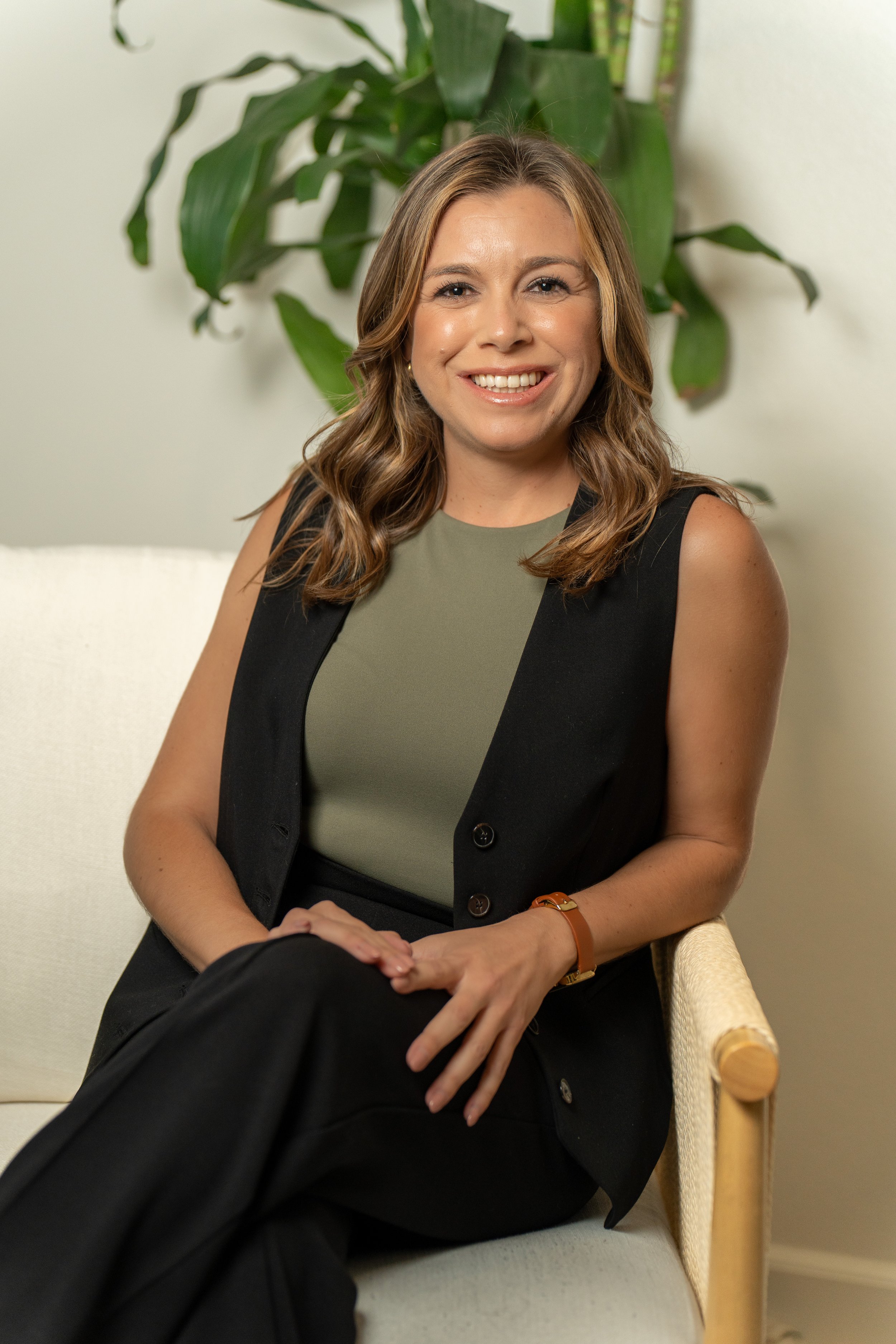A woman with wavy light brown hair smiling, sitting on a beige chair with wooden armrests, wearing a sleeveless black vest over a green top, with a large green leafy plant behind her, in an indoor setting.