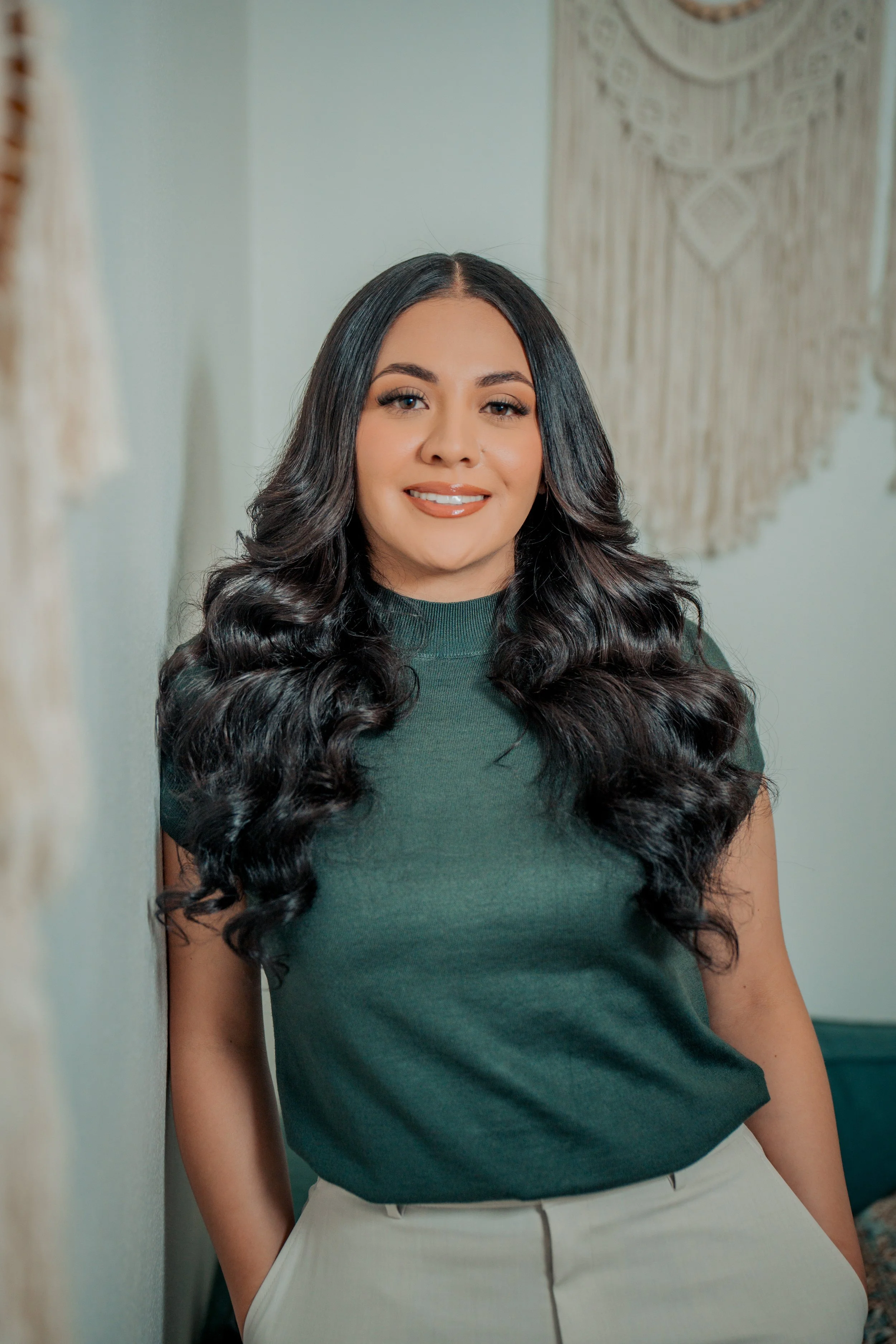 A woman with long, dark, wavy hair smiling, wearing a green top and cream-colored pants, standing indoors with a woven wall hanging in the background.