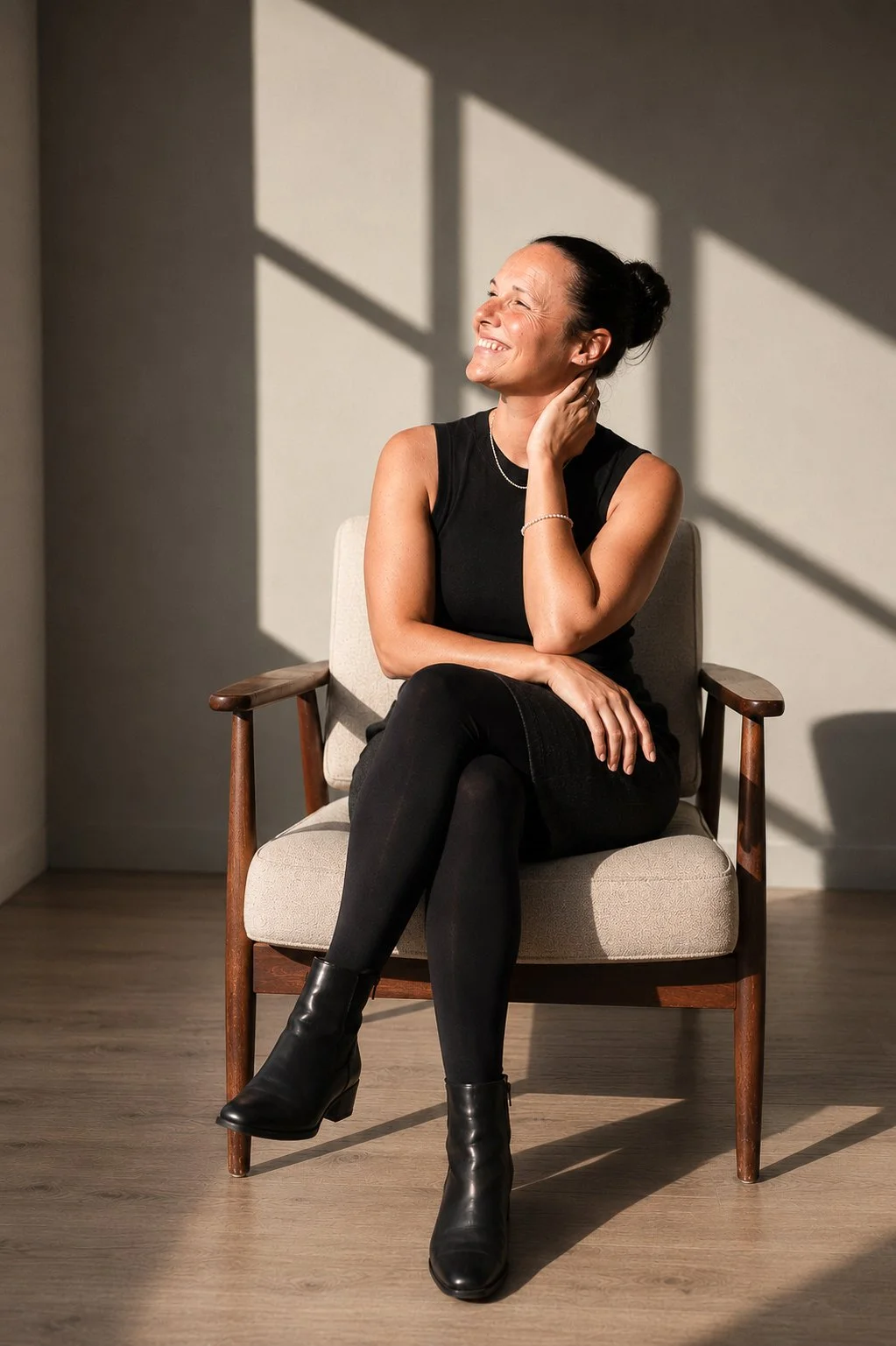 Lächelnde Frau mit hochgesteckten dunklen Haaren und schwarzem ärmellosem Oberteil sitzt entspannt in einem Sessel und blickt zur Seite. Warmes Sonnenlicht wirft geometrische Schatten auf die helle Wand.