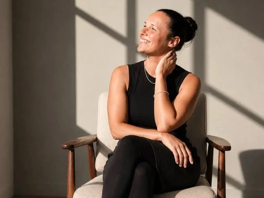 Lächelnde Frau mit hochgesteckten dunklen Haaren und schwarzem ärmellosem Oberteil sitzt entspannt in einem Sessel und blickt zur Seite. Warmes Sonnenlicht wirft geometrische Schatten auf die helle Wand.