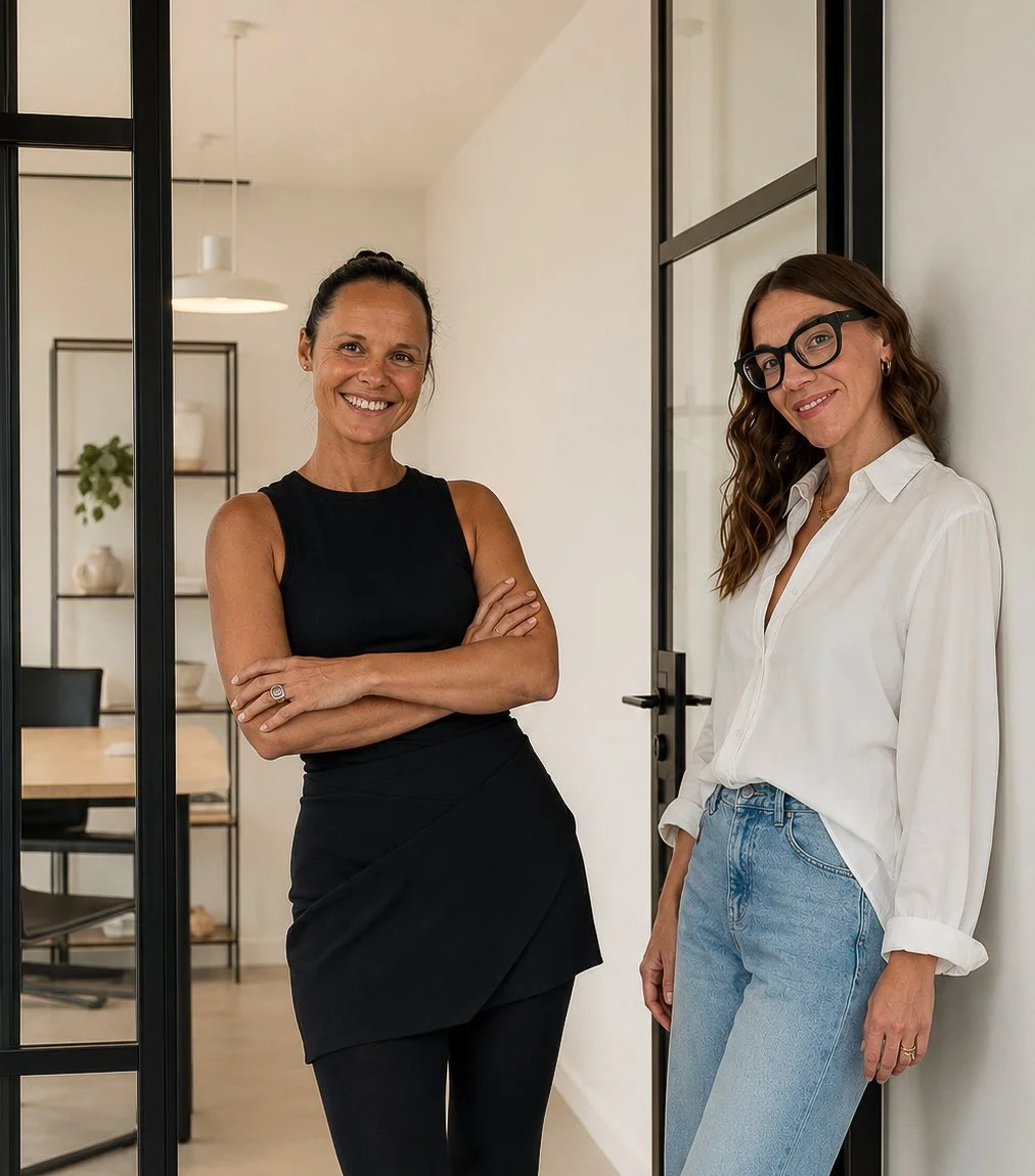 Zwei lächelnde Frauen stehen in einem hellen, modern eingerichteten Büro mit schwarzen Glasrahmen. Eine trägt ein schwarzes ärmelloses Outfit, die andere eine weiße Bluse, Jeans und eine schwarze Brille.
