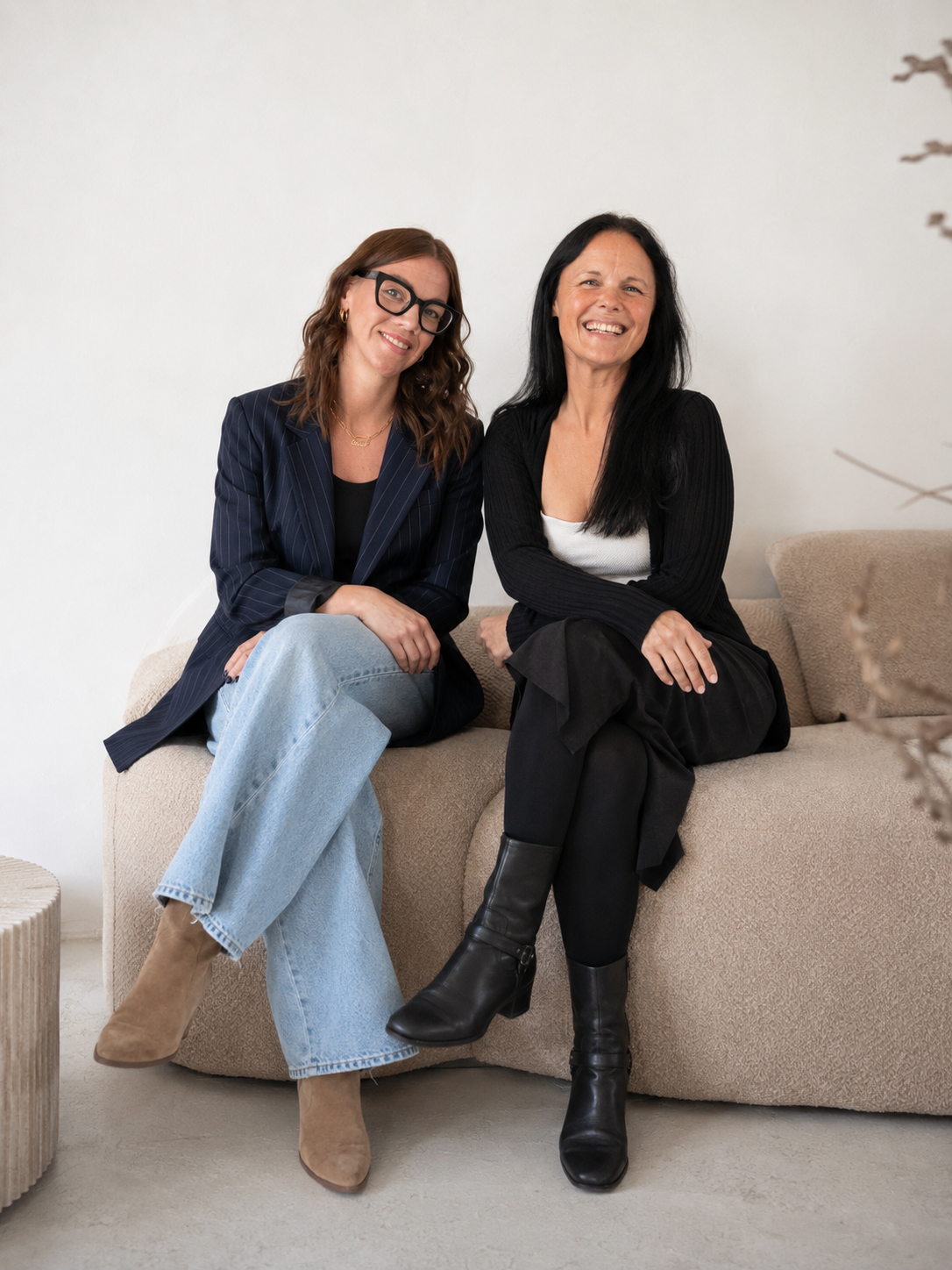 Zwei lächelnde Frauen sitzen nebeneinander auf einem beigen Sofa vor einer hellen Wand. Eine trägt eine dunkle Nadelstreifenjacke, helle Jeans und eine schwarze Brille, die andere ein schwarzes Outfit mit weißem Oberteil.