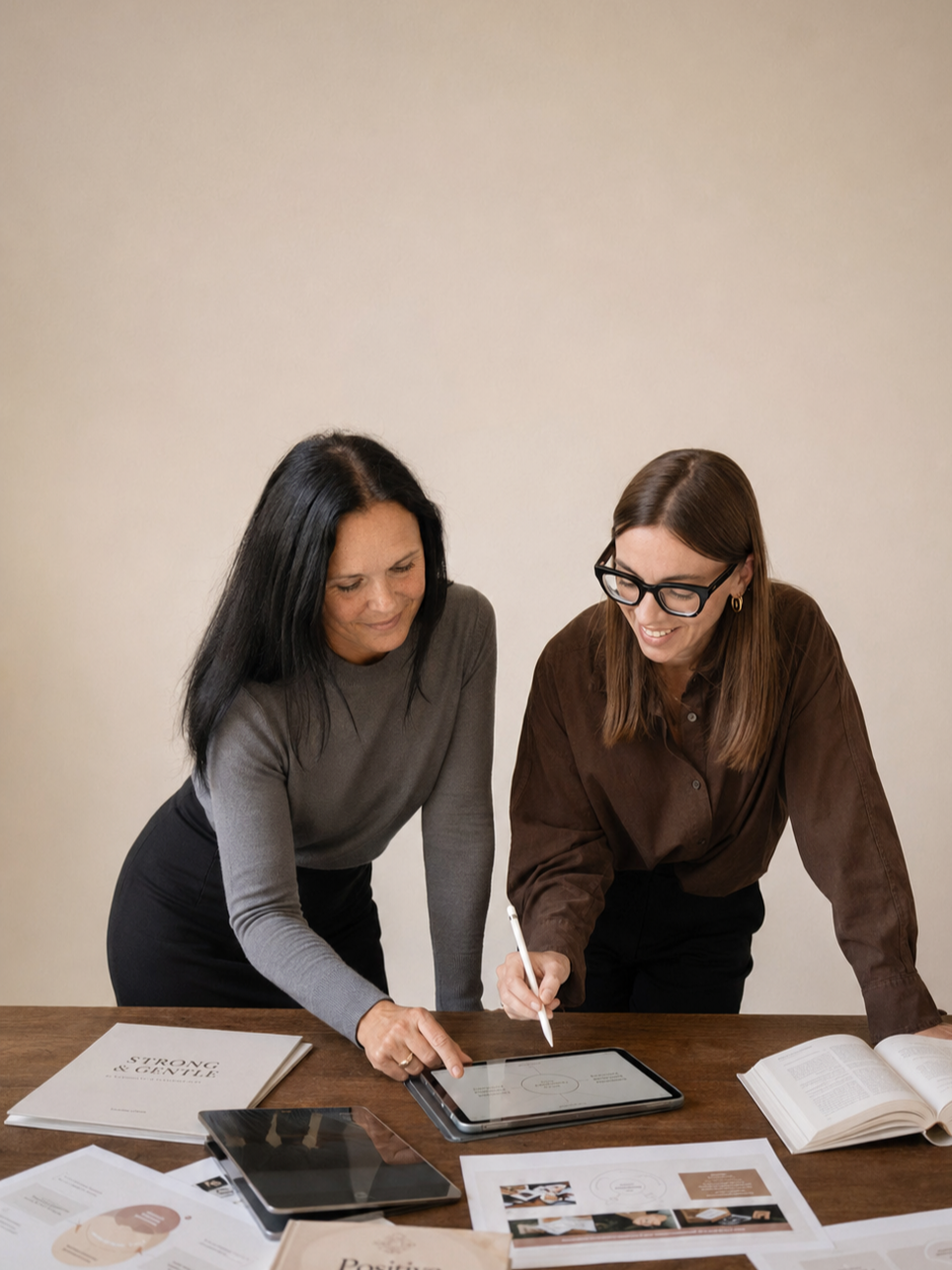 Zwei Frauen stehen gemeinsam an einem Holztisch und arbeiten konzentriert an einem Tablet mit Eingabestift. Auf dem Tisch liegen Unterlagen und Arbeitsmaterialien.