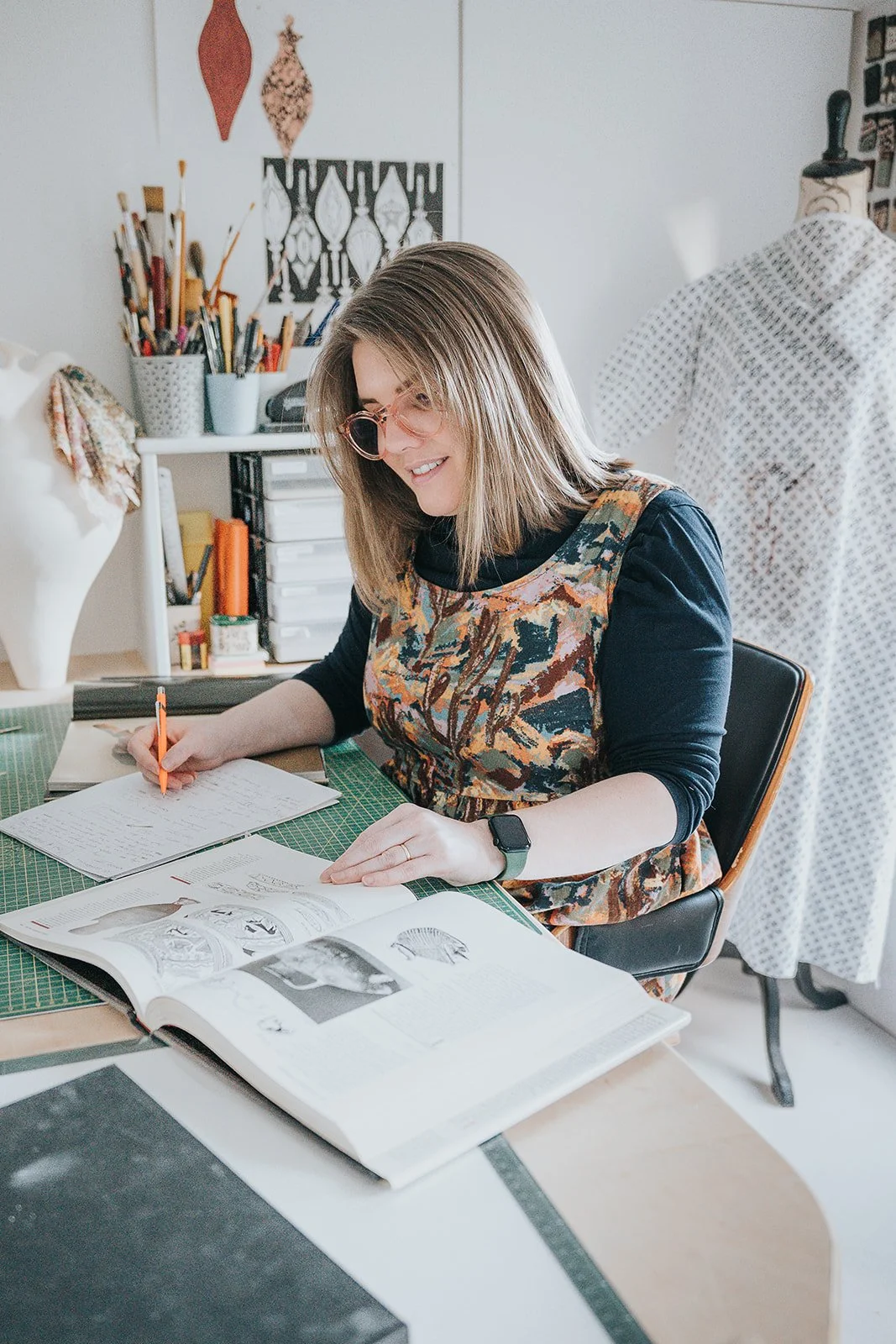 A woman with shoulder-length hair and glasses, sitting at a desk, writing in a notebook and looking at an open art book, in an art studio with art supplies and a mannequin in the background.
