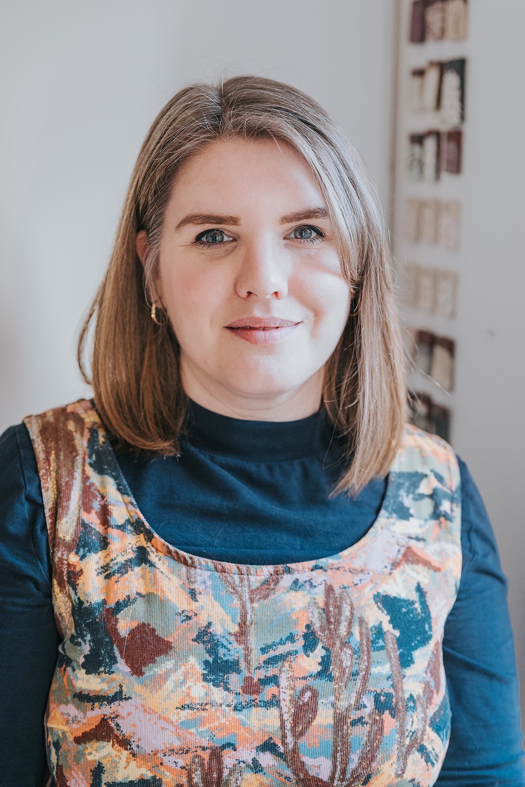 A woman with shoulder-length blonde hair and green eyes smiling softly, wearing a navy top with a colourful, patterned dungarees, standing indoors with a blurred background.