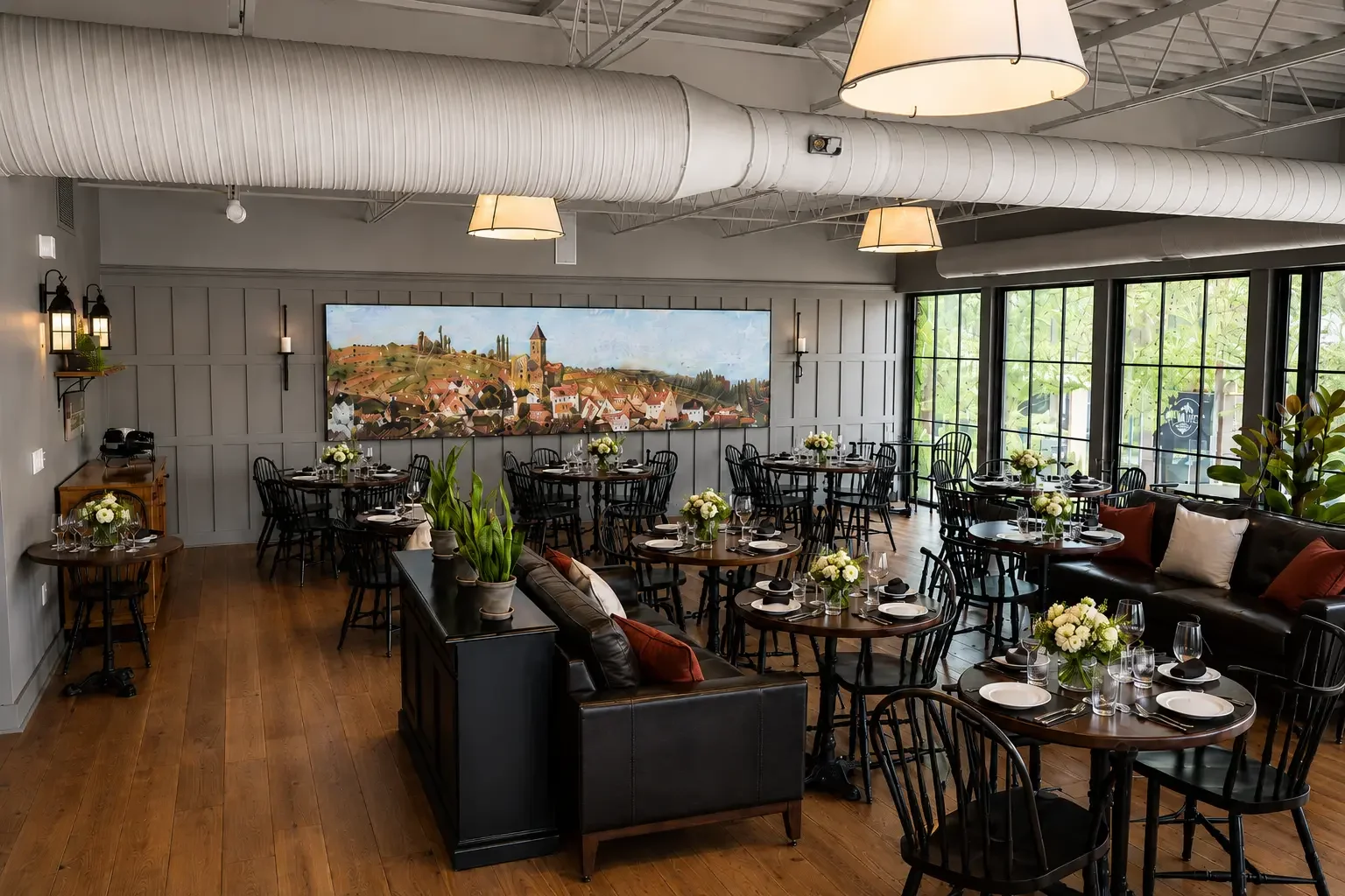 A restaurant dining area with wooden tables and black chairs, decorated with white flower centerpieces. Large windows on one side let in natural light. Artwork of a village scene hangs on the back wall, and there is a black leather couch with pillows and potted plants. The ceiling has exposed ducts and hanging lights.