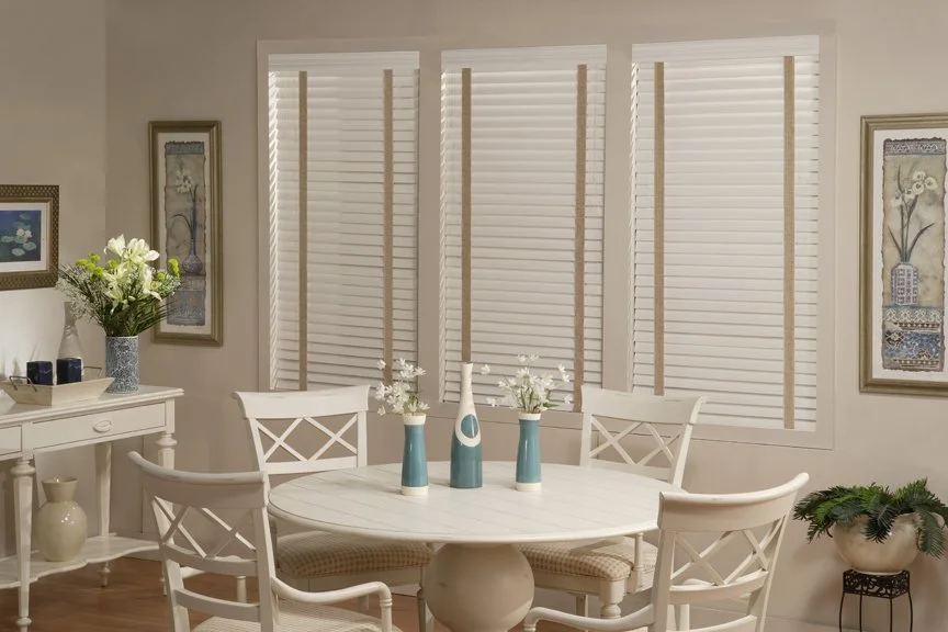 A white dining table with four matching chairs, decorated with three vases of white flowers, in a bright room with large windows covered with white blinds, framed artwork on the walls, and floral arrangements and decor on nearby furniture.