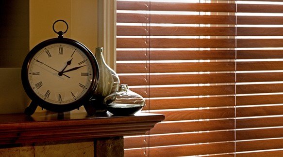 A black alarm clock and decorative vases on a wooden mantel near a window with closed wooden blinds.