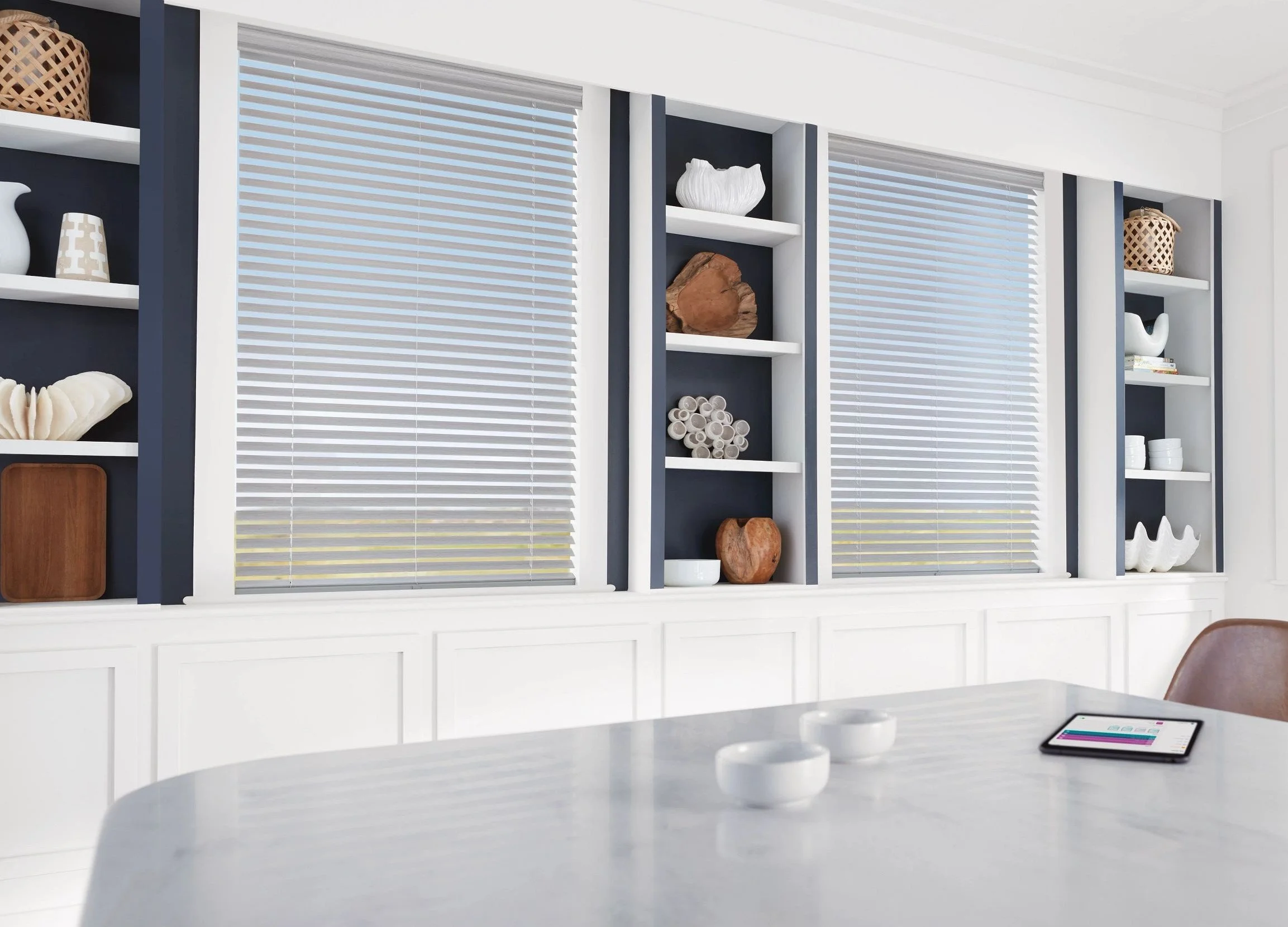 A bright dining room with a white table, a tablet, and decorative bowls. Behind, shelves with white and brown decorative objects and closed blinds on windows.