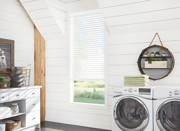 Laundry room with white shiplap walls, a window with blinds, a front-loading washer and dryer, a round wall shelf with plants, folded towels, and a white storage cabinet.