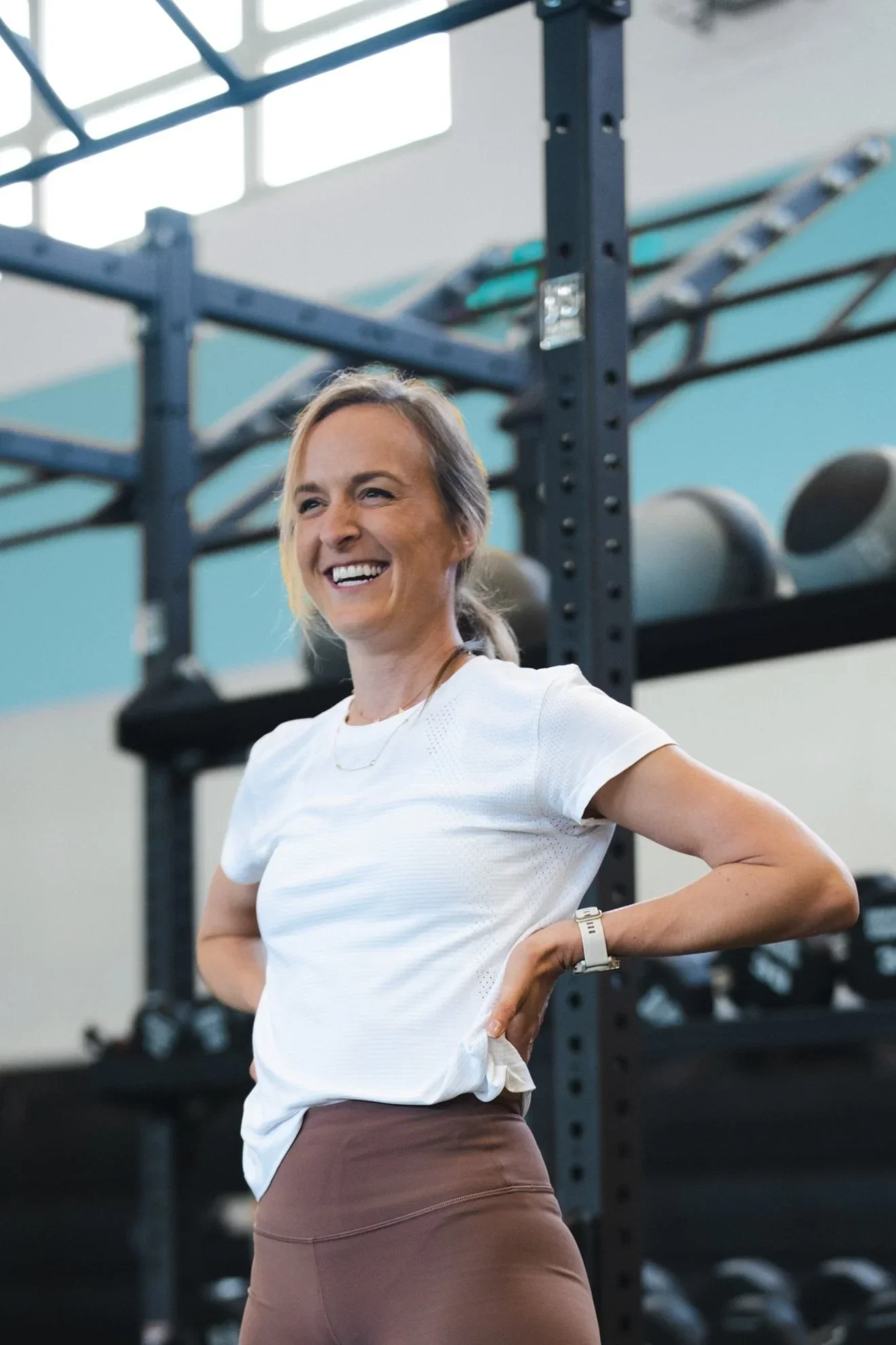 A woman in workout attire smiling in a gym with black shelving and exercise equipment in the background