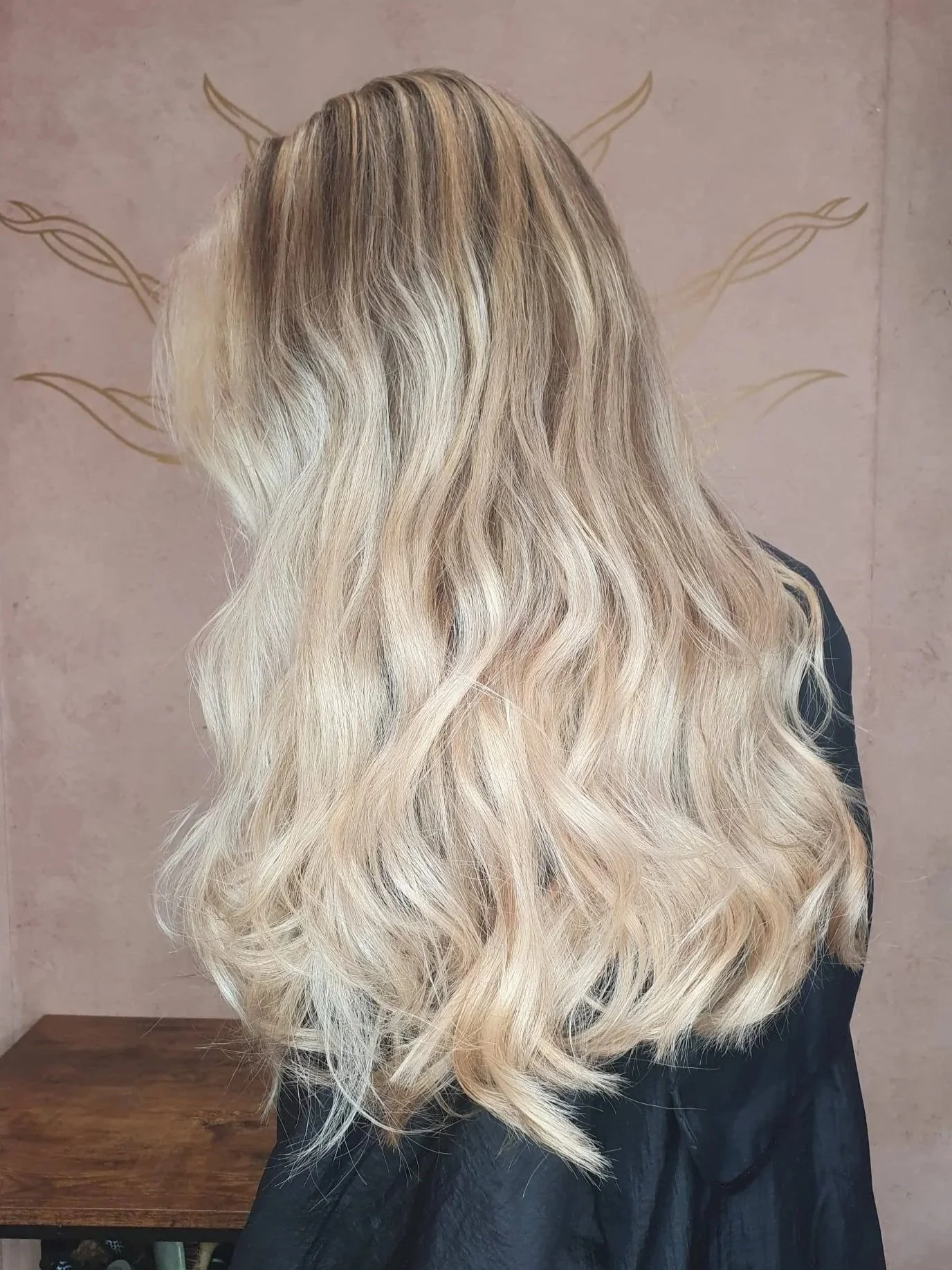 Back view of a woman with long, wavy, blonde hair standing indoors against a wall with a decorative pattern.