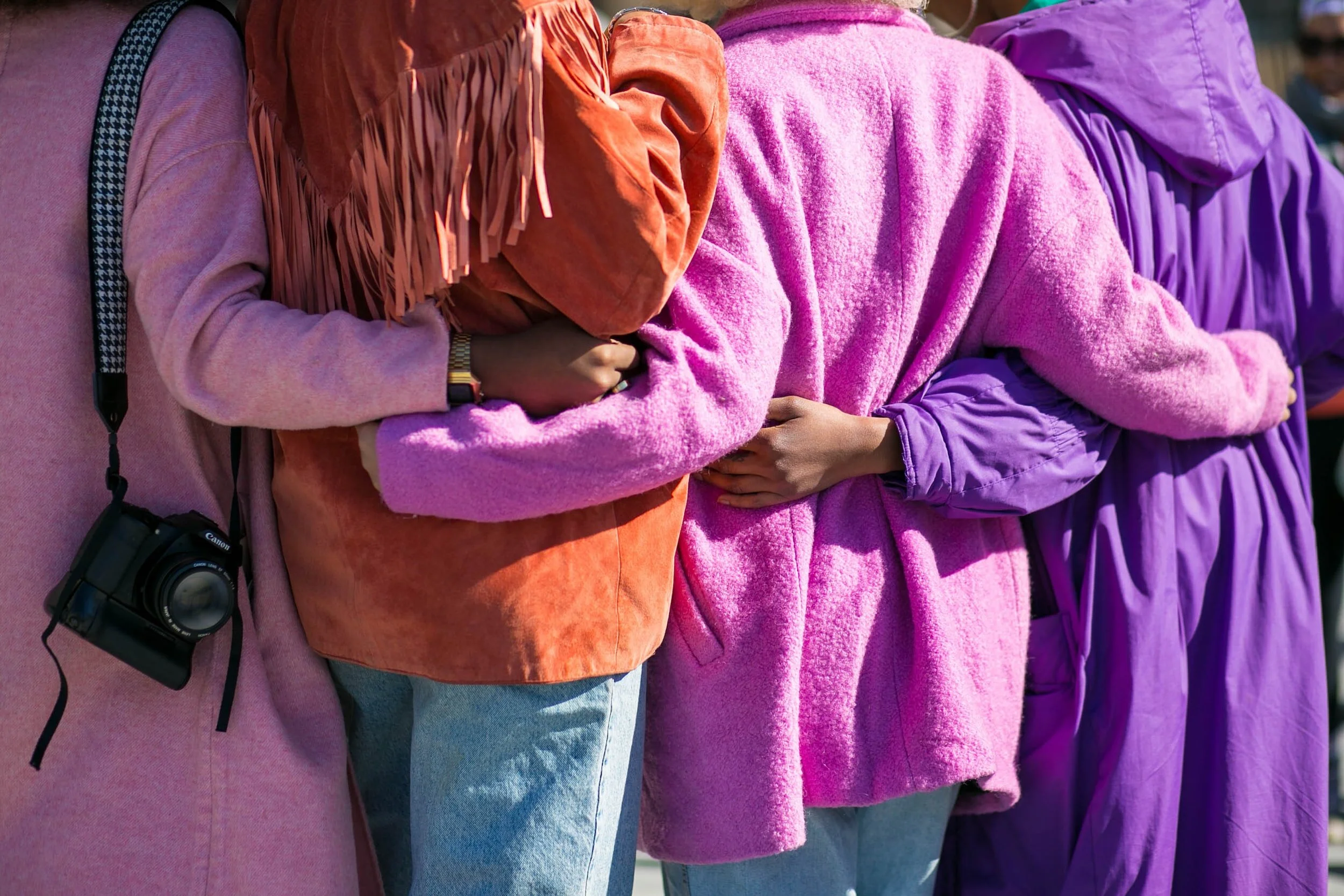 A group of people wearing colorful jackets in orange, pink, and purple wrap their arms around one another's backs