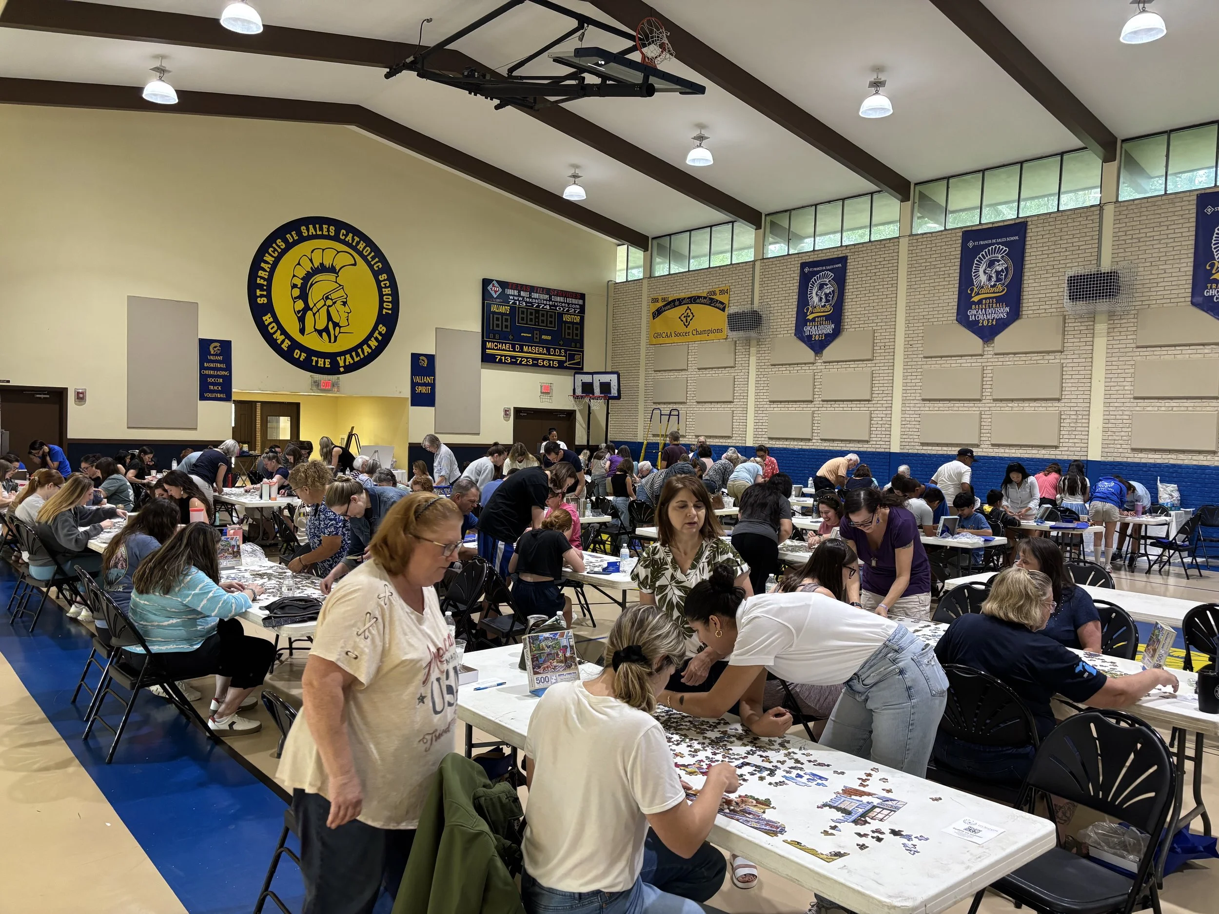 People gathering in a gymnasium for a puzzle competition, with tables filled with puzzle pieces and boxes. The gym has banners, scoreboard, and basketball hoop visible.