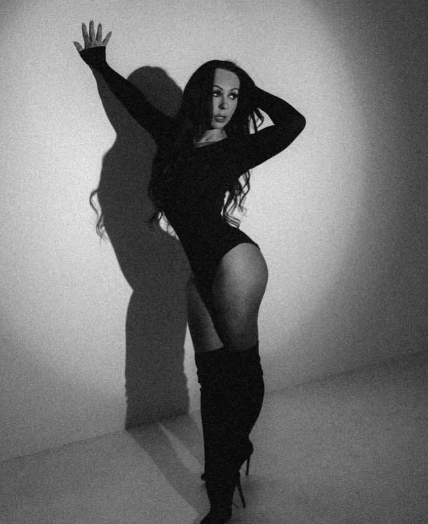 A woman with long, wavy hair in a dimly lit environment, wearing a strapless dress. The background has blue and purple lights, suggesting a party or event setting.