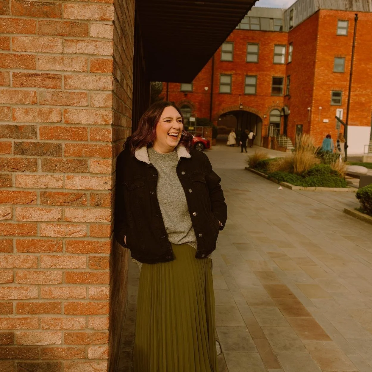 A woman with shoulder-length dark hair, wearing a black jacket and a green pleated skirt, is smiling and leaning against a brick wall in an outdoor urban area during daytime, interested in wellness.