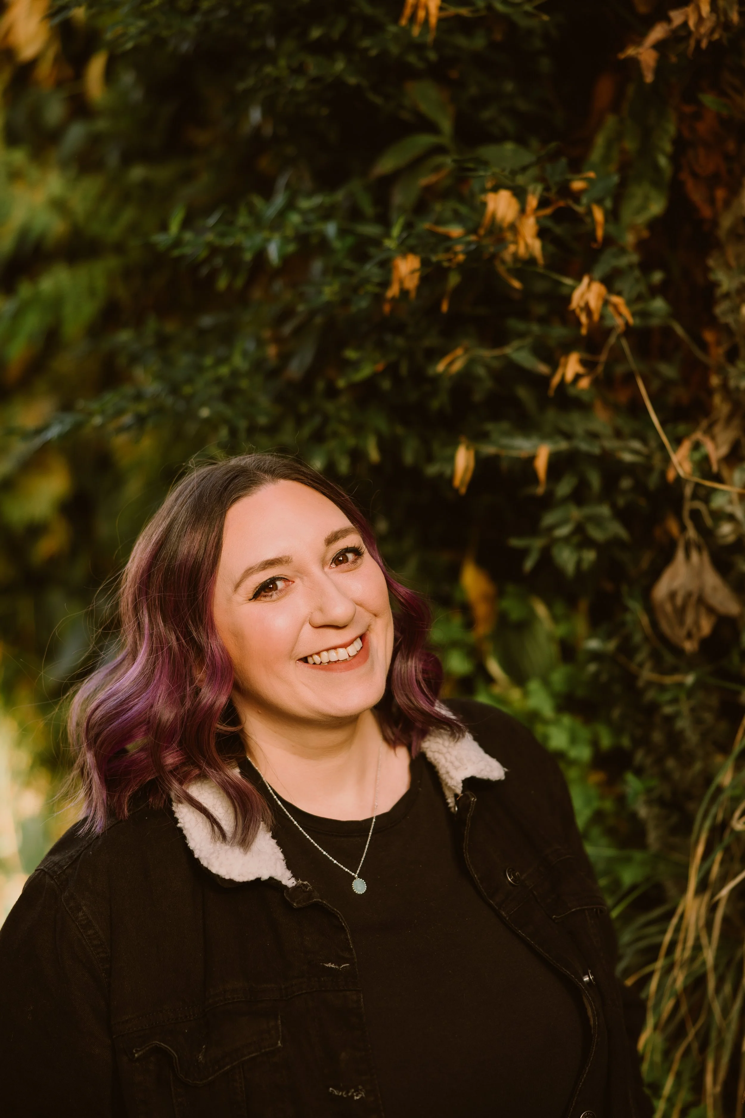 A young woman with shoulder-length, wavy, purple-tinted hair and light skin, smiling at a camera. She is wearing a black jacket with a cream-colored fleece lining and a silver necklace with a round pendant. The background is filled with green and yellow foliage.