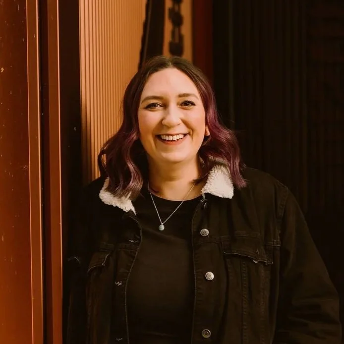 A woman with shoulder-length purple hair, smiling, wearing a black jacket with a furry collar and a necklace, standing indoors near a wooden panel and dark background.
