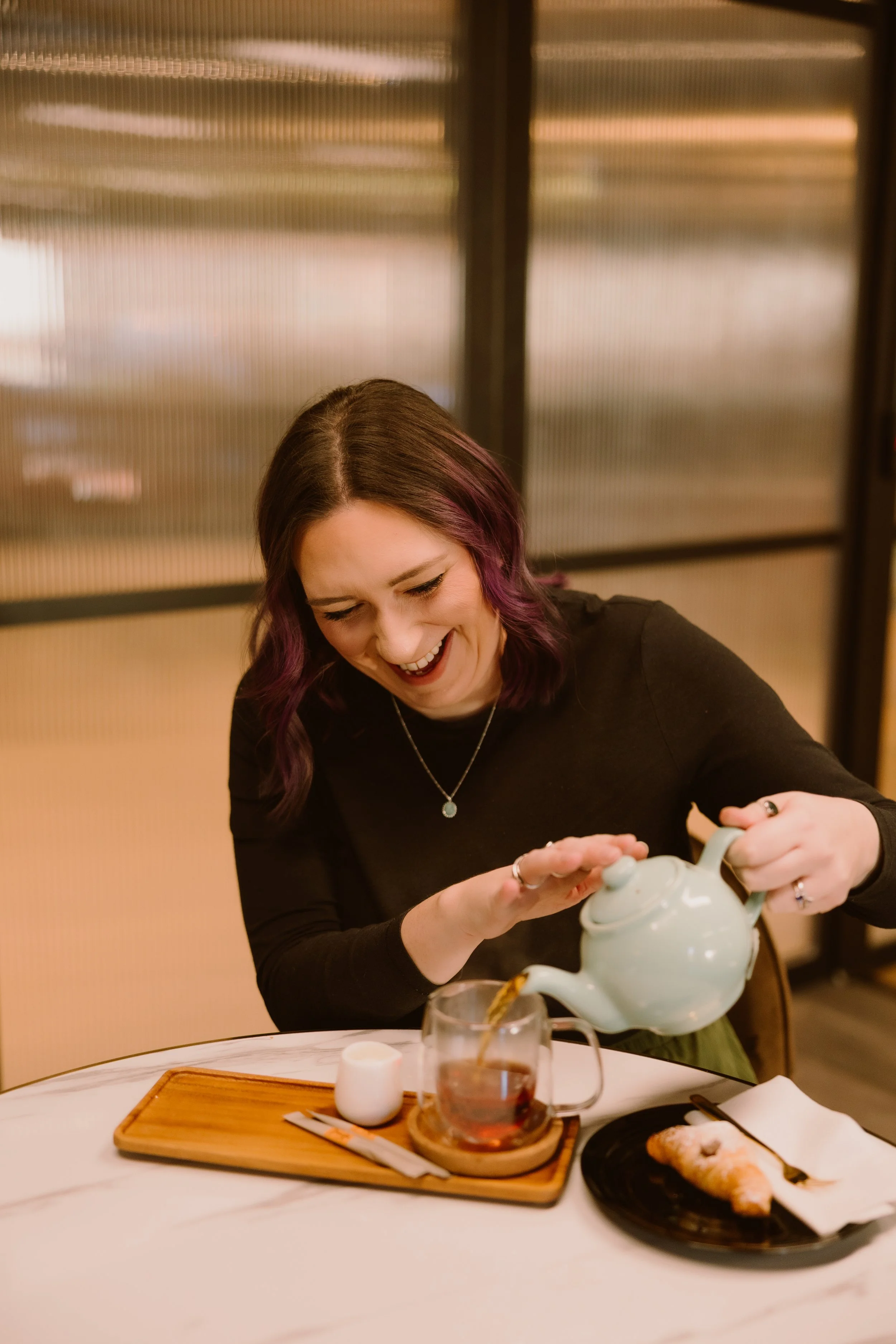 A woman with purple hair is pouring tea into a glass placed on a table with breakfast food and a wooden tray.