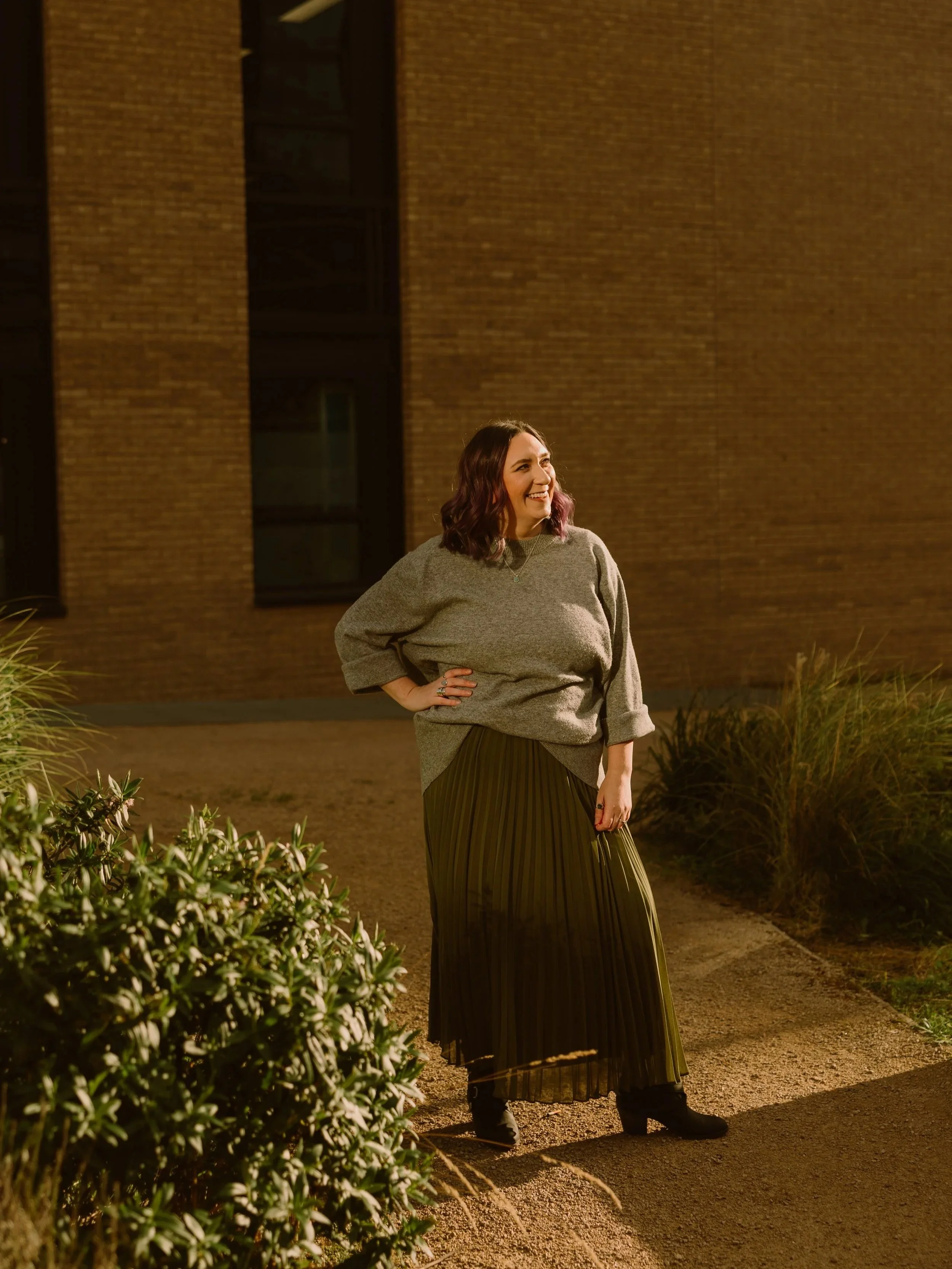 A woman with shoulder-length wavy hair, standing outdoors at night, smiling, wearing a gray sweater and a long pleated skirt, with plants in the foreground and a brick building in the background.