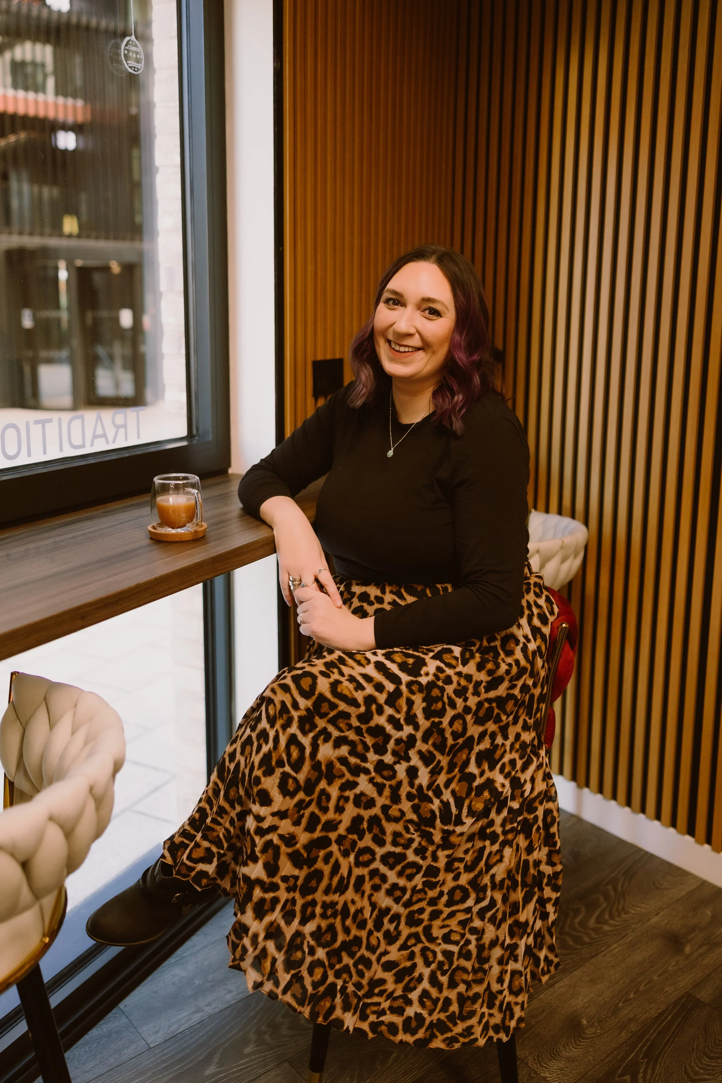 A woman with shoulder-length, wavy hair with purple tips, sitting inside a cafe near a window, wearing a black top and a leopard print skirt, smiling at the camera with a glass of coffee on the counter beside her.