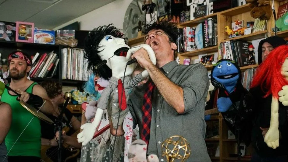 A man is laughing and holding a puppet with black hair, red lips, and a gray suit, while a person in the background plays drums at a puppet show event. Several puppets and books are visible on shelves.